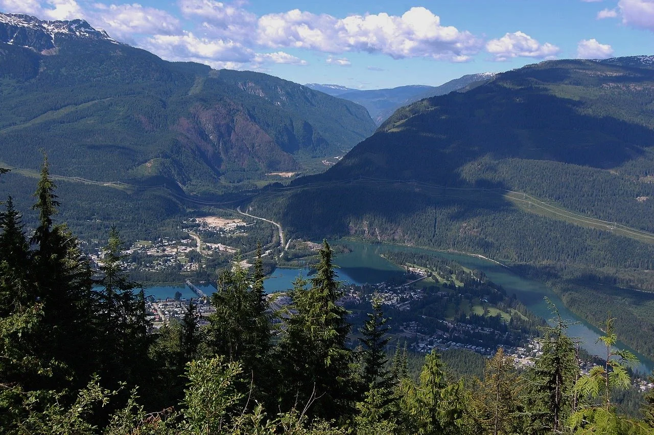 Panoramic view of a town nestled in a valley surrounded by mountains, with a river running through the town and evergreen trees in the foreground.