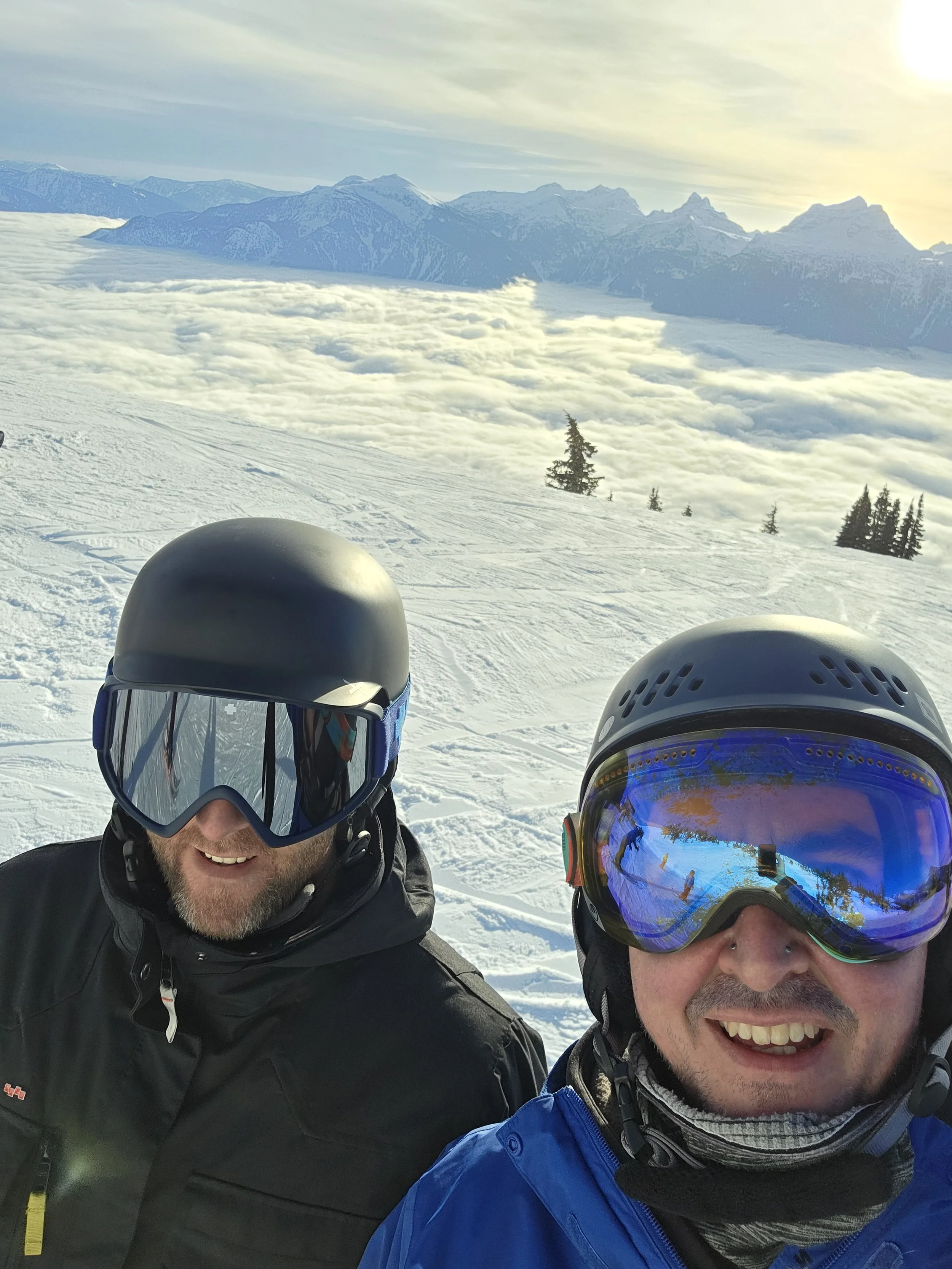 Two men wearing snow helmets and goggles taking a selfie on a snowy mountain with snow-covered peaks and a layer of clouds in the background.