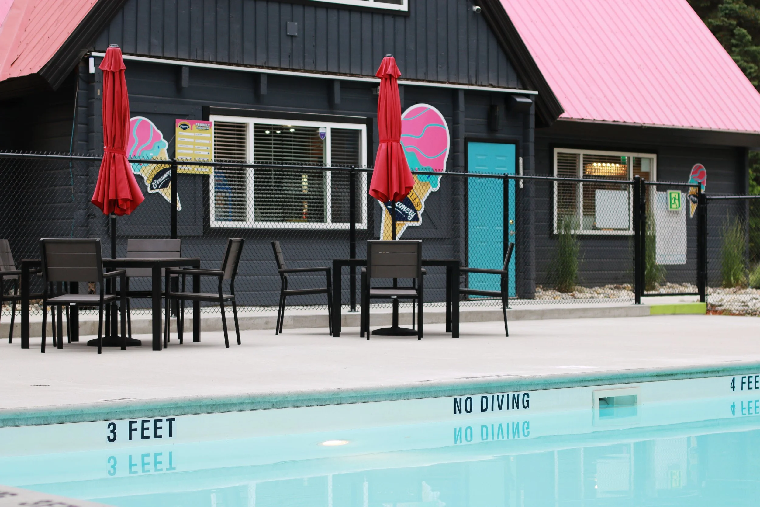 An outdoor swimming pool with a water depth sign that reads "NO DIVING" and "3 FEET," surrounded by black patio chairs, red umbrellas, and a black building with colorful ice cream cone murals on the exterior.