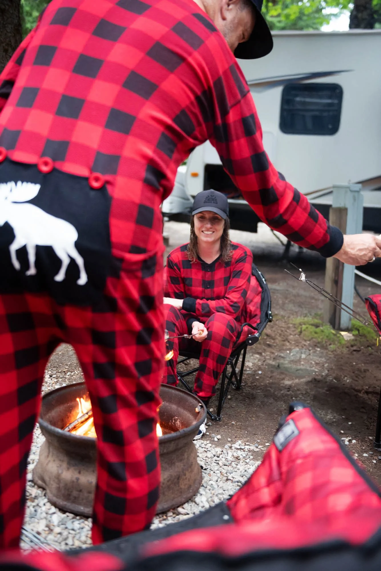 People in red and black plaid clothing around a campfire with a woman sitting in a chair smiling.