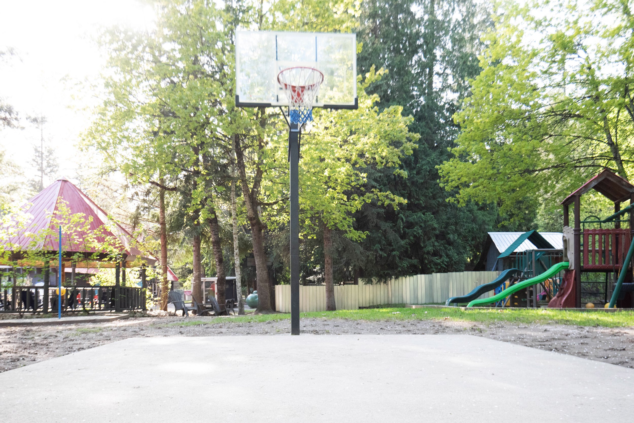 A playground with a basketball hoop in the foreground, surrounded by trees and playground structures, with a red roof and slides in the background.