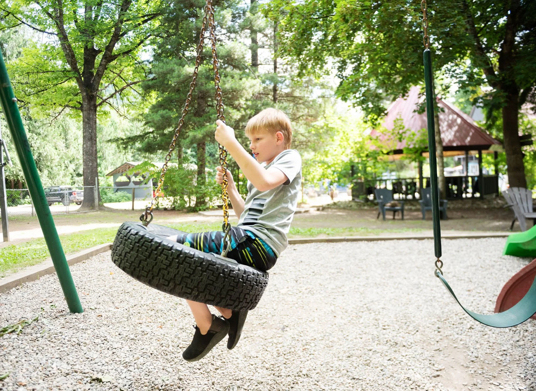 A young boy with blonde hair playing on a tire swing in a park filled with lush green trees. The tire is attached to a swing set with chains, and the boy is seated on the swing, holding onto the chains with a focused expression. In the background, th