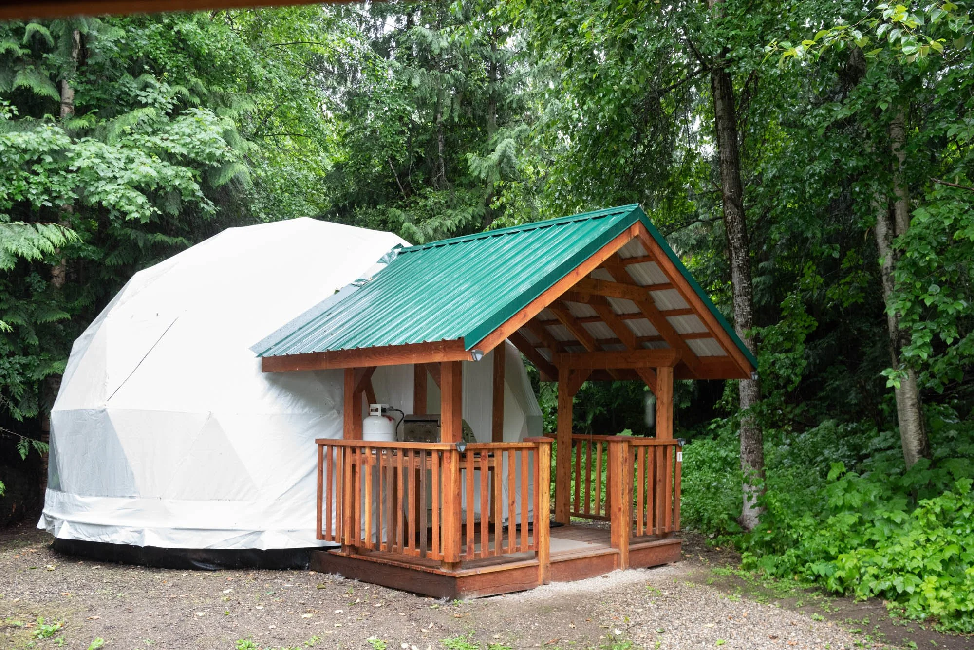 A small wooden cabin with a green metal roof, partly enclosed and built on a wooden deck, situated in a lush green forest. The cabin has a propane tank and other items inside, visible through an open side.