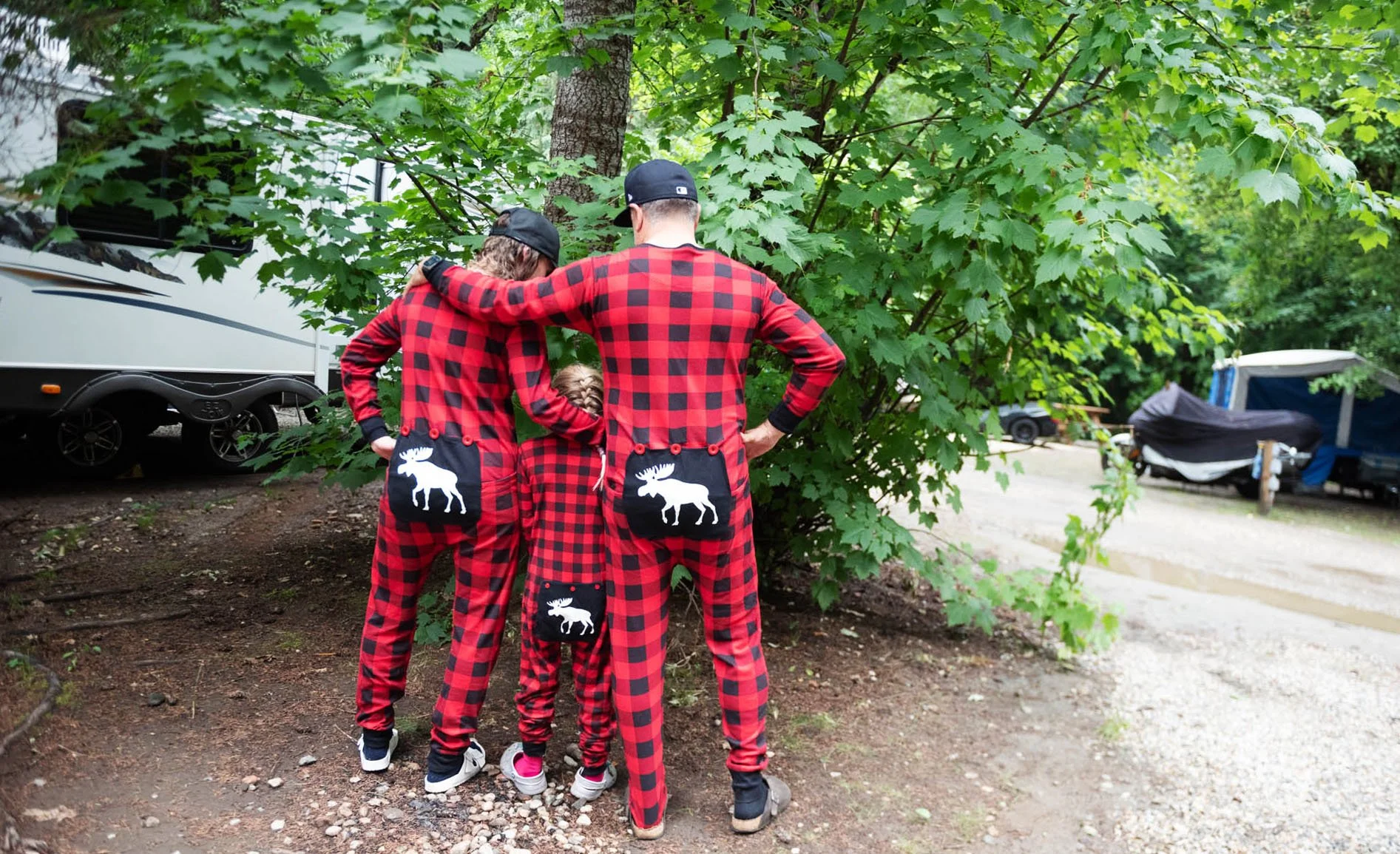 Three people wearing matching red and black plaid pajamas with moose patches on the back, standing close together with arms around each other in a wooded area near RVs and trees.