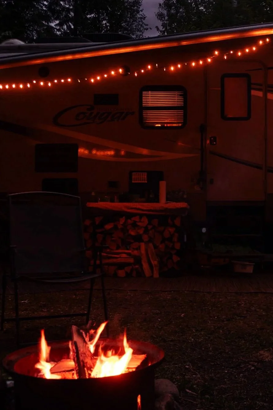 A campfire with flames burning in a fire pit, a stack of chopped firewood behind a camper trailer, and string lights hanging from the top of the trailer at dusk or nighttime.