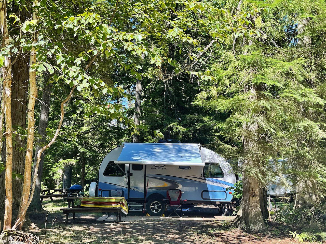 A blue and white camper trailer parked in a forested area with trees and green foliage, a picnic table with a colorful tablecloth, and camping chairs.