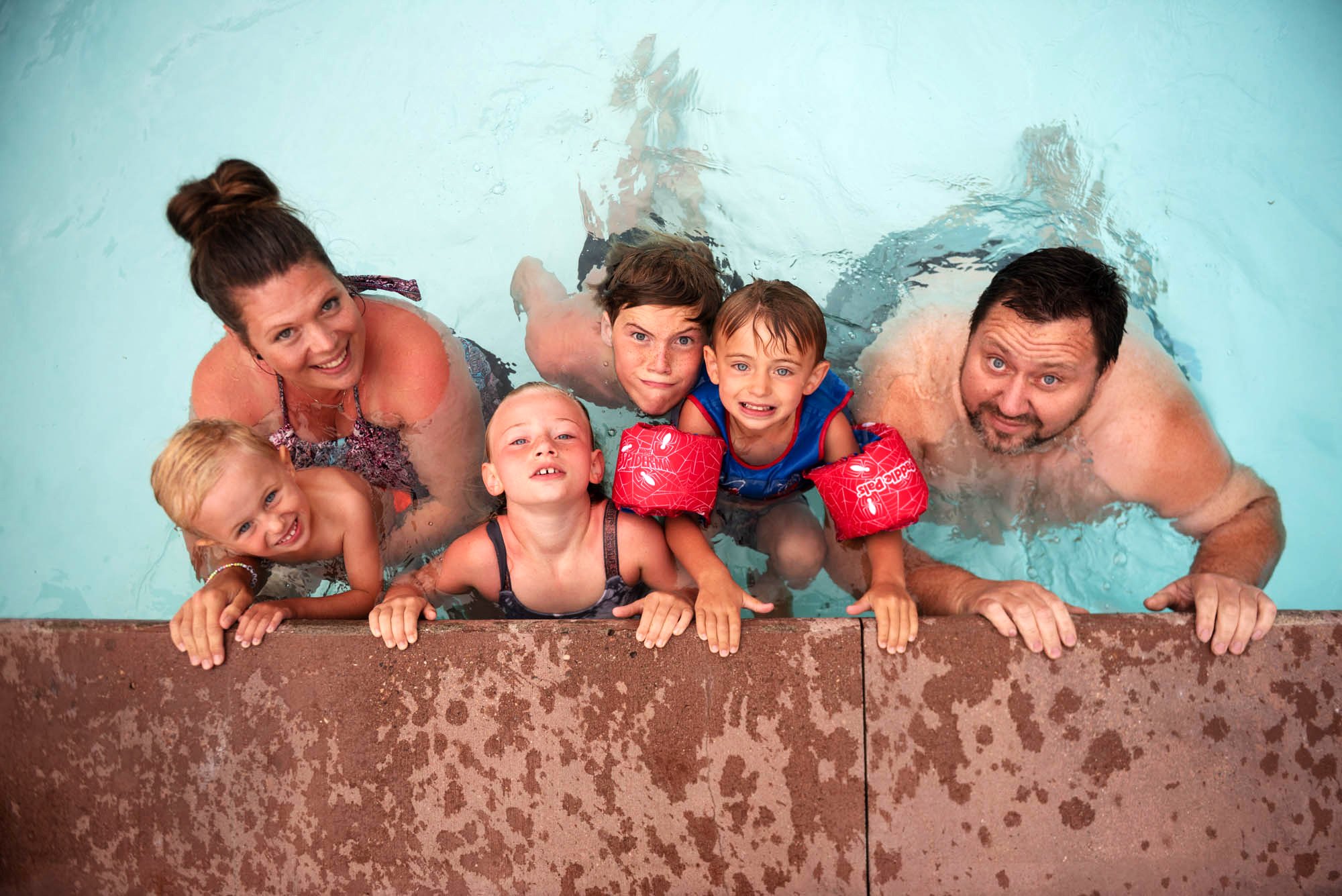 A family of six in a swimming pool, smiling and looking up at the camera, with their arms resting on the pool edge.