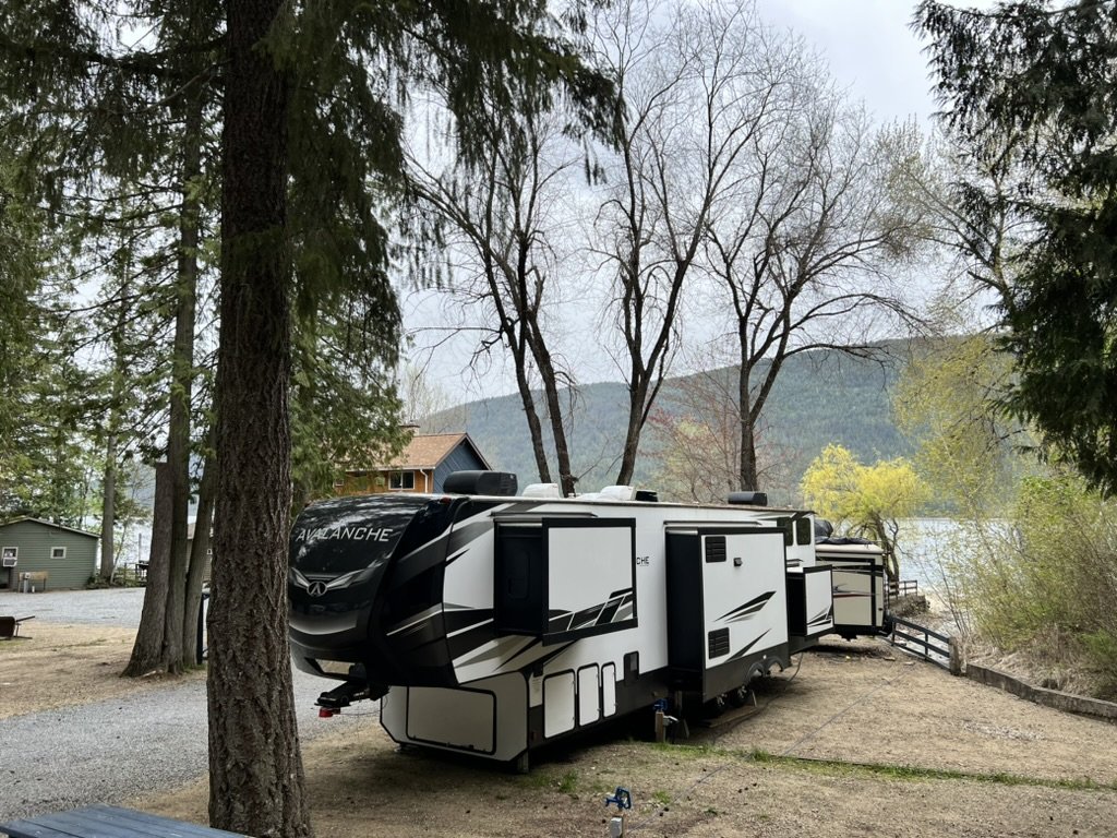 A white travel trailer with black accents parked among trees near a lake, with mountains and a wooden house in the background.