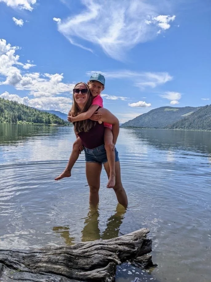 A woman in sunglasses holding a young girl on her back while standing in a lake with a scenic mountain and cloudy sky in the background.
