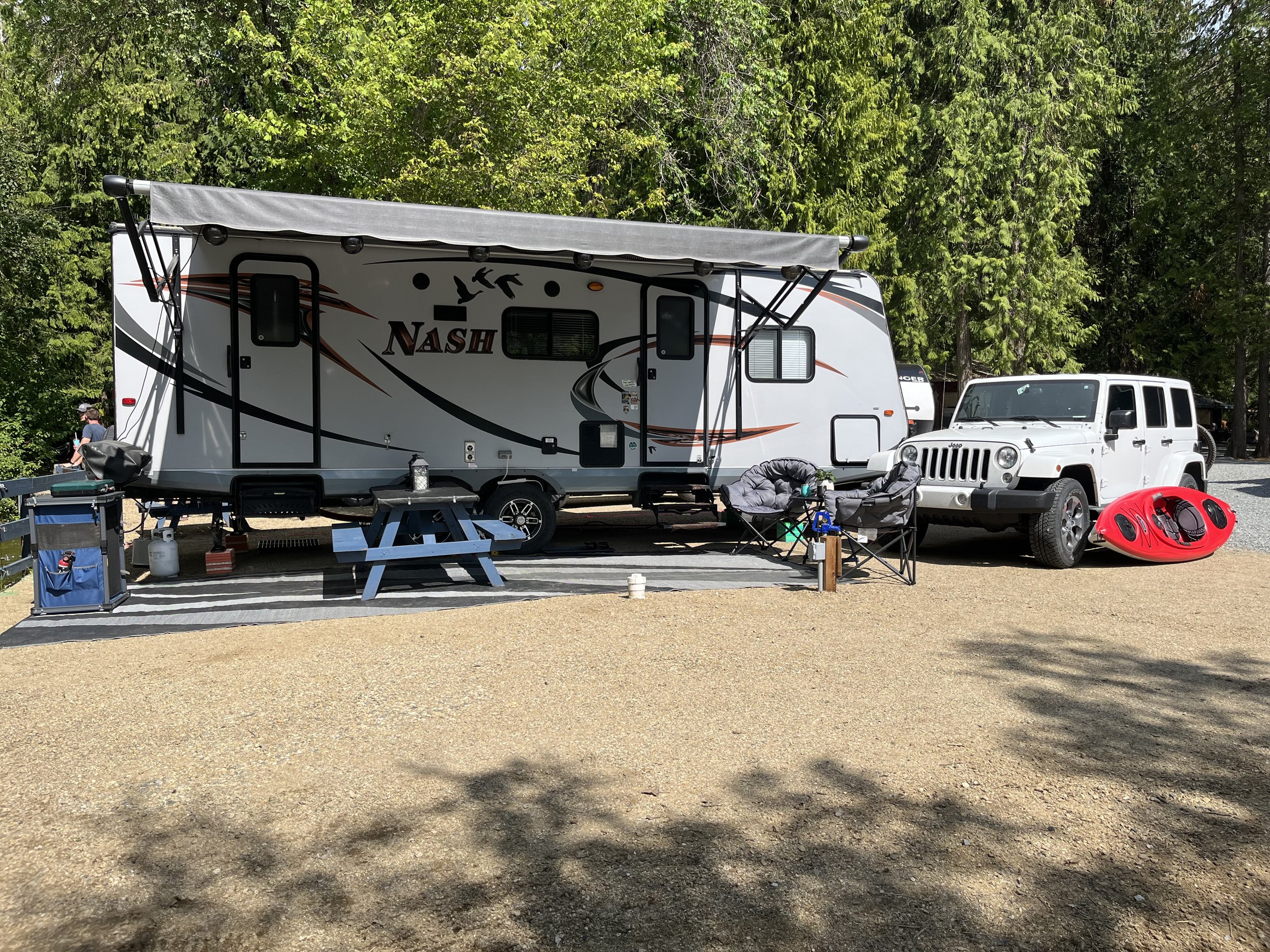 A white Nash travel trailer with black and orange graphics parked on a gravel lot, surrounded by green trees, with camping gear and a white Jeep nearby.