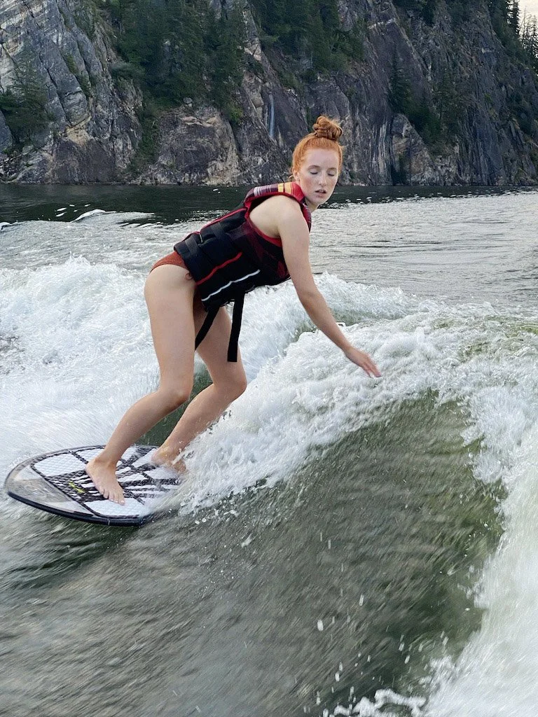 A young woman with red hair and a bun is surfing on a wakeboard in a lake, wearing a life vest near rocky cliffs and trees.