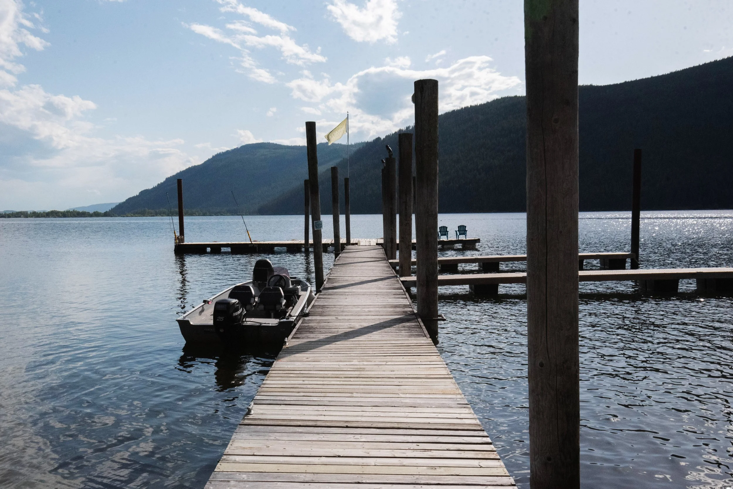 A wooden pier extending into a calm lake with two chairs at the end, surrounded by mountains and a partly cloudy sky.