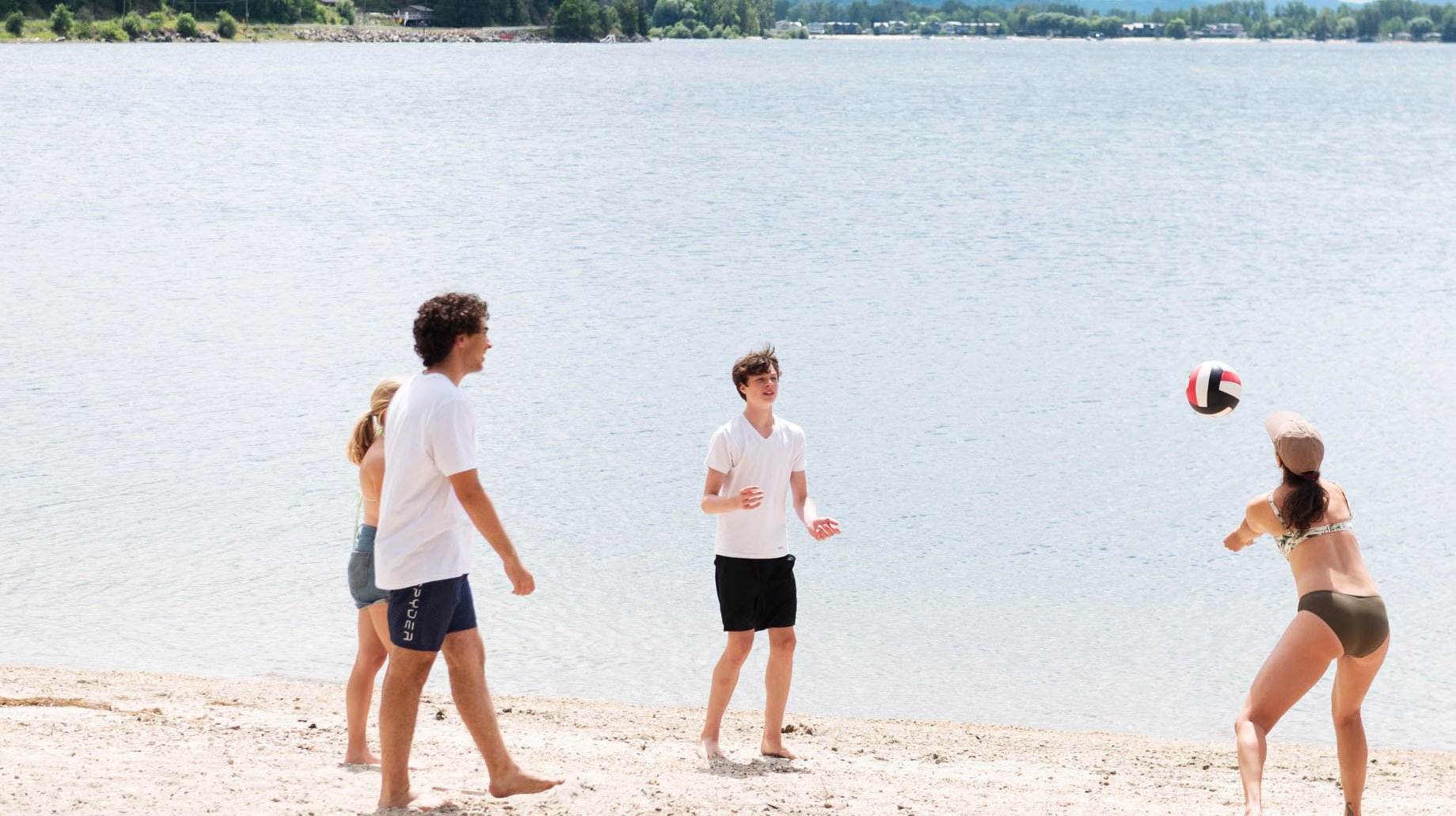 Group of five people playing volleyball on a sandy beach near a body of water, with trees in the background. The woman on the right is about to hit the volleyball, while the others watch.