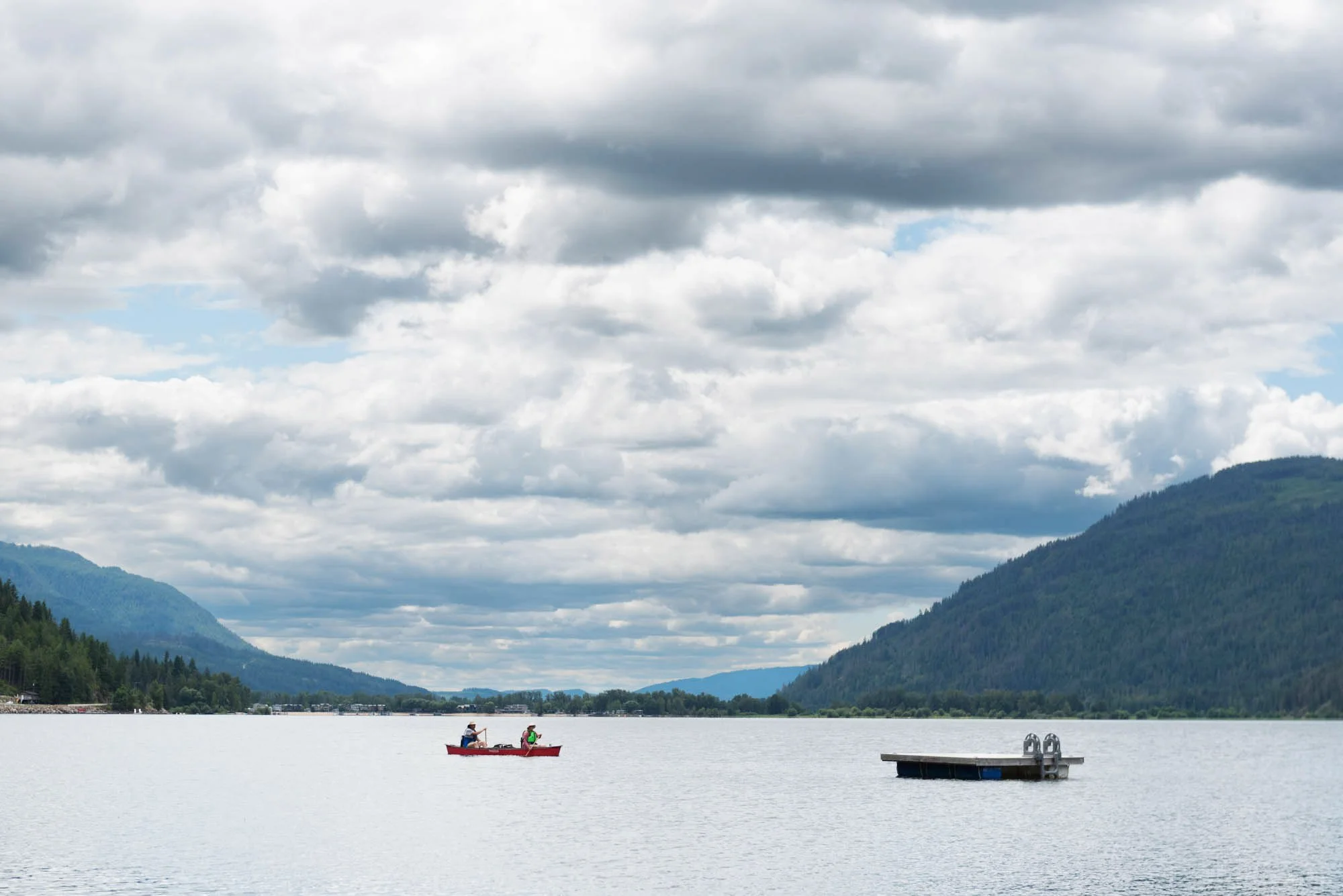Two people kayaking on a lake surrounded by forested mountains with partly cloudy sky.