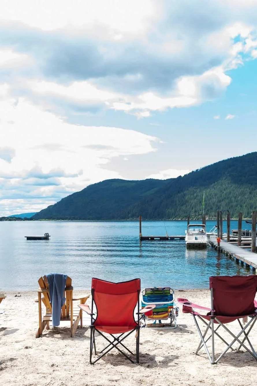 Beach scene with four lawn chairs on sandy shore, overlooking a calm lake with boats and a wooden dock, surrounded by green mountains under a partly cloudy sky.