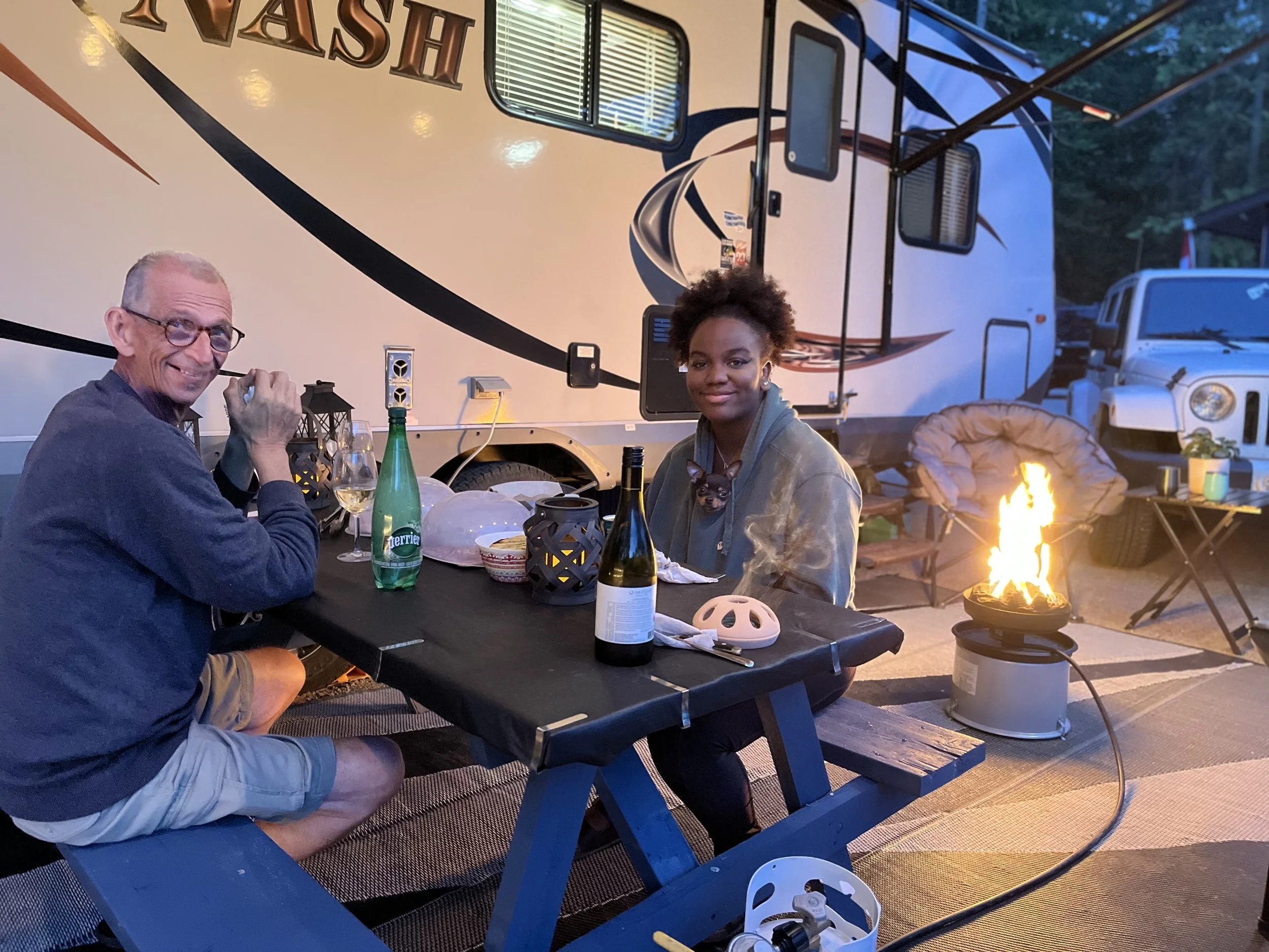Two people sitting at a picnic table outdoors in the evening, with a fire in a fire pit, bottles, and glasses on the table, and an RV in the background.