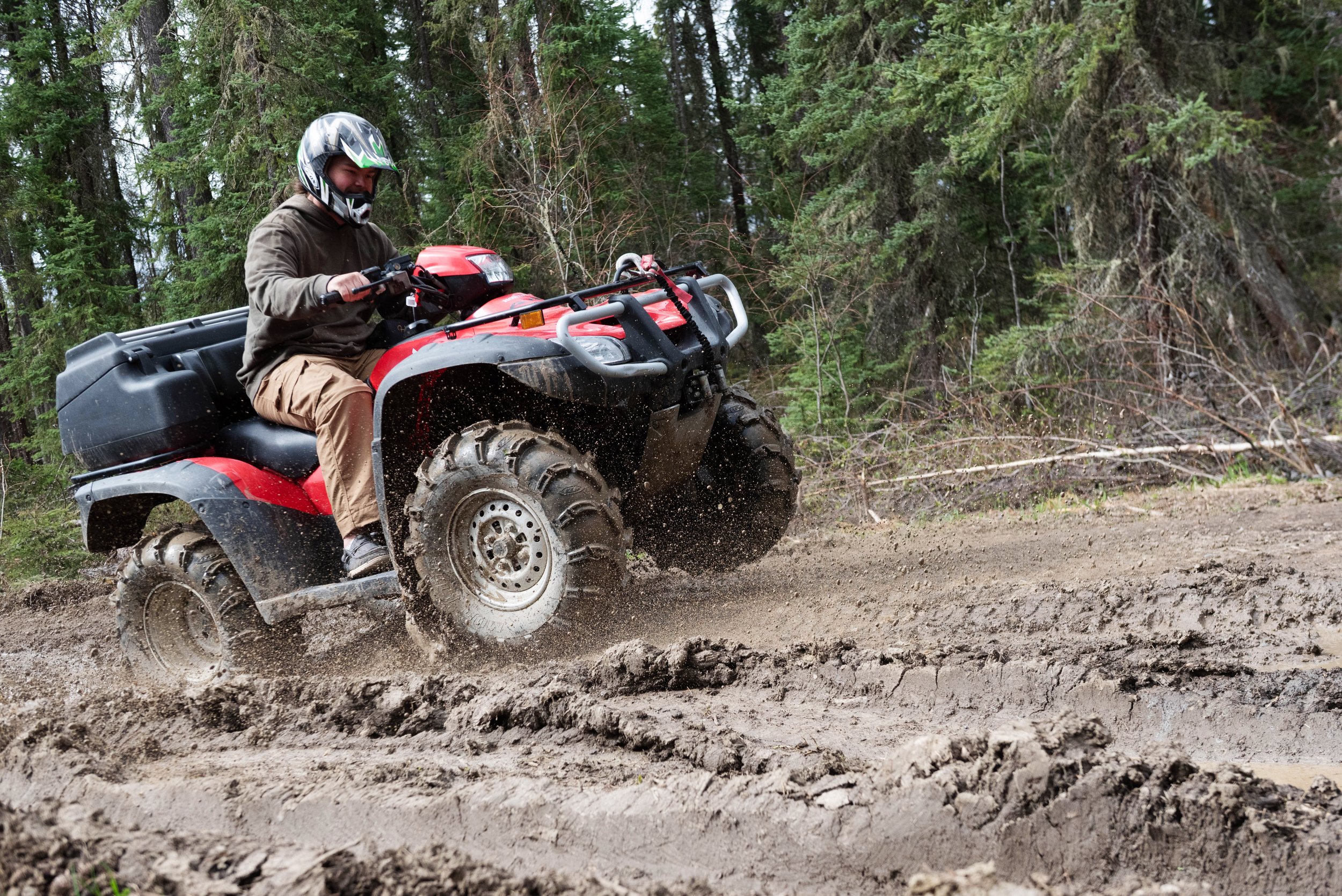 A person wearing a helmet and brown jacket riding a red ATV through a dirt trail in a forest with green trees.