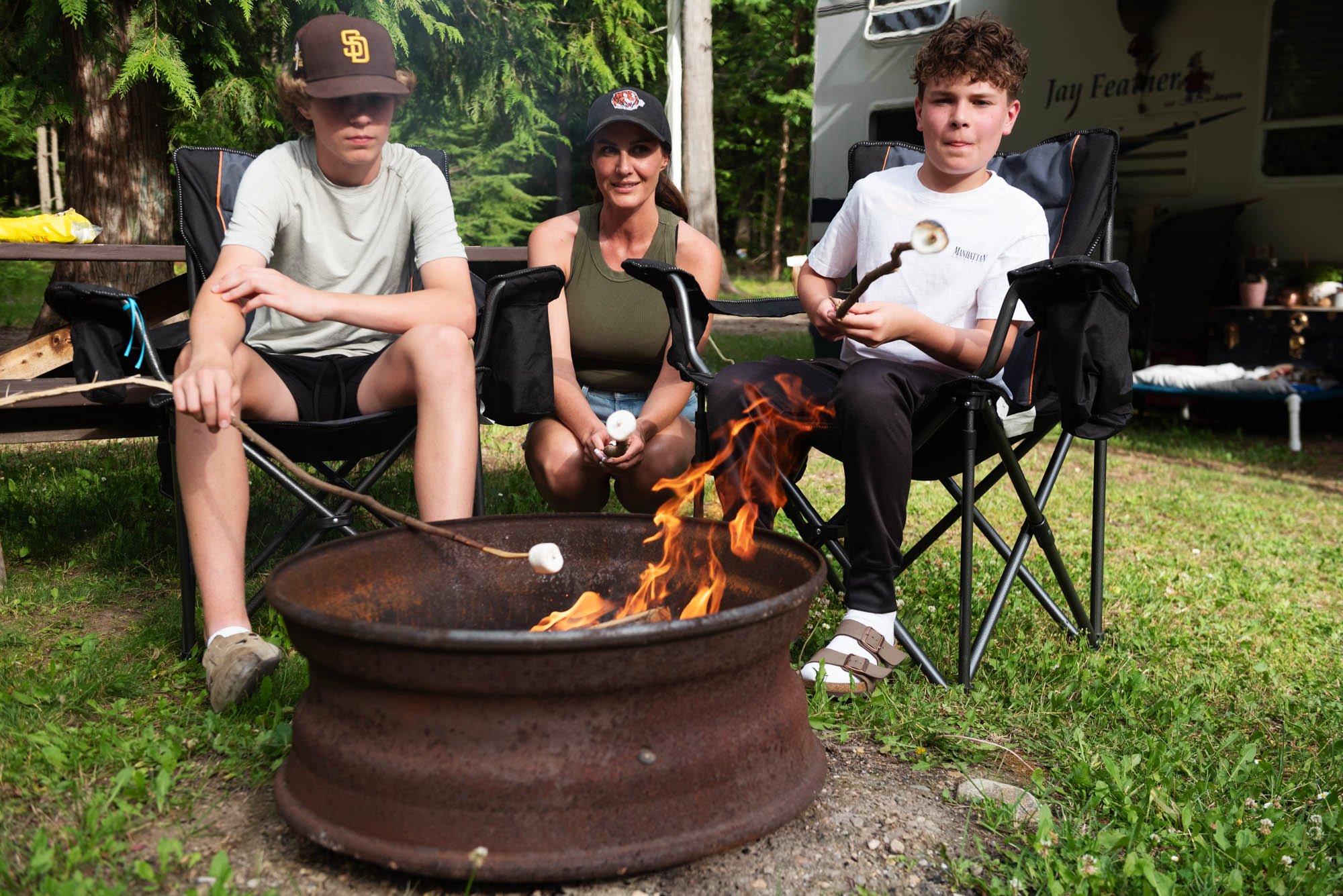 Three people sitting outdoors near a campfire in a wooded area, with tents and trees in the background. Two boys and one woman, all wearing casual clothing including caps, are seated in camping chairs. The boy on the right is holding a roasting stick with marshmallows over the fire.