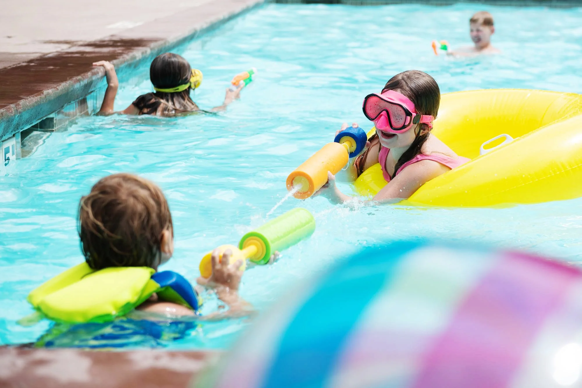 Children in a swimming pool with floatation devices and water guns, engaging in a swimming lesson or play.