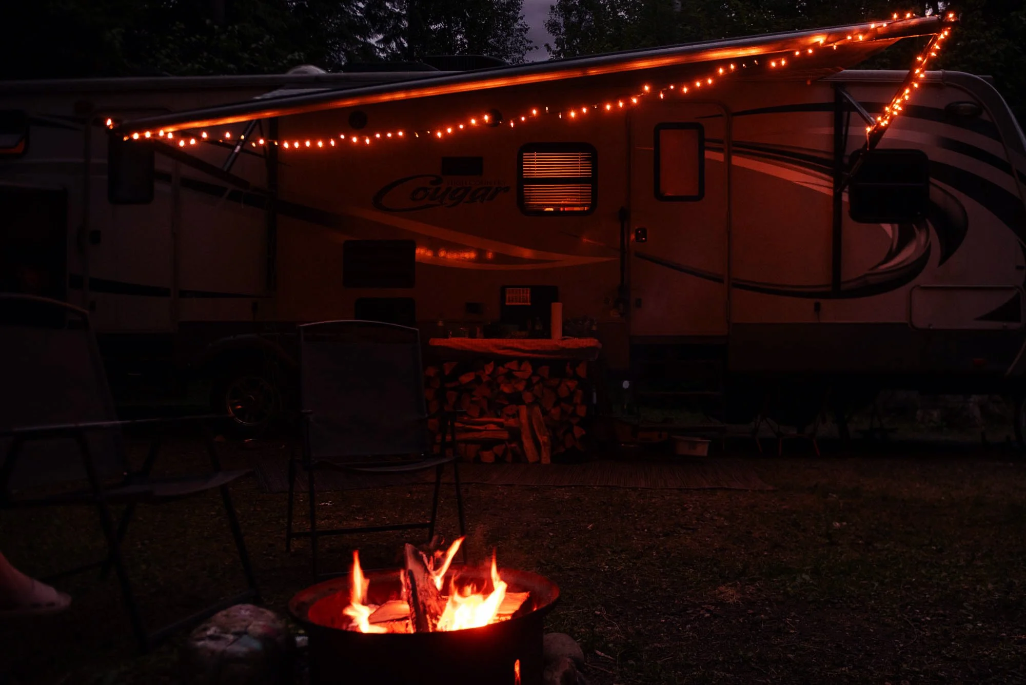 A recreational vehicle with string lights hung along the top edge, parked outdoors at night. In front of the RV, there is a fire pit with a small controlled fire burning, surrounded by outdoor chairs and tables. The scene suggests a camping or outdoor gathering setting at dusk or night.