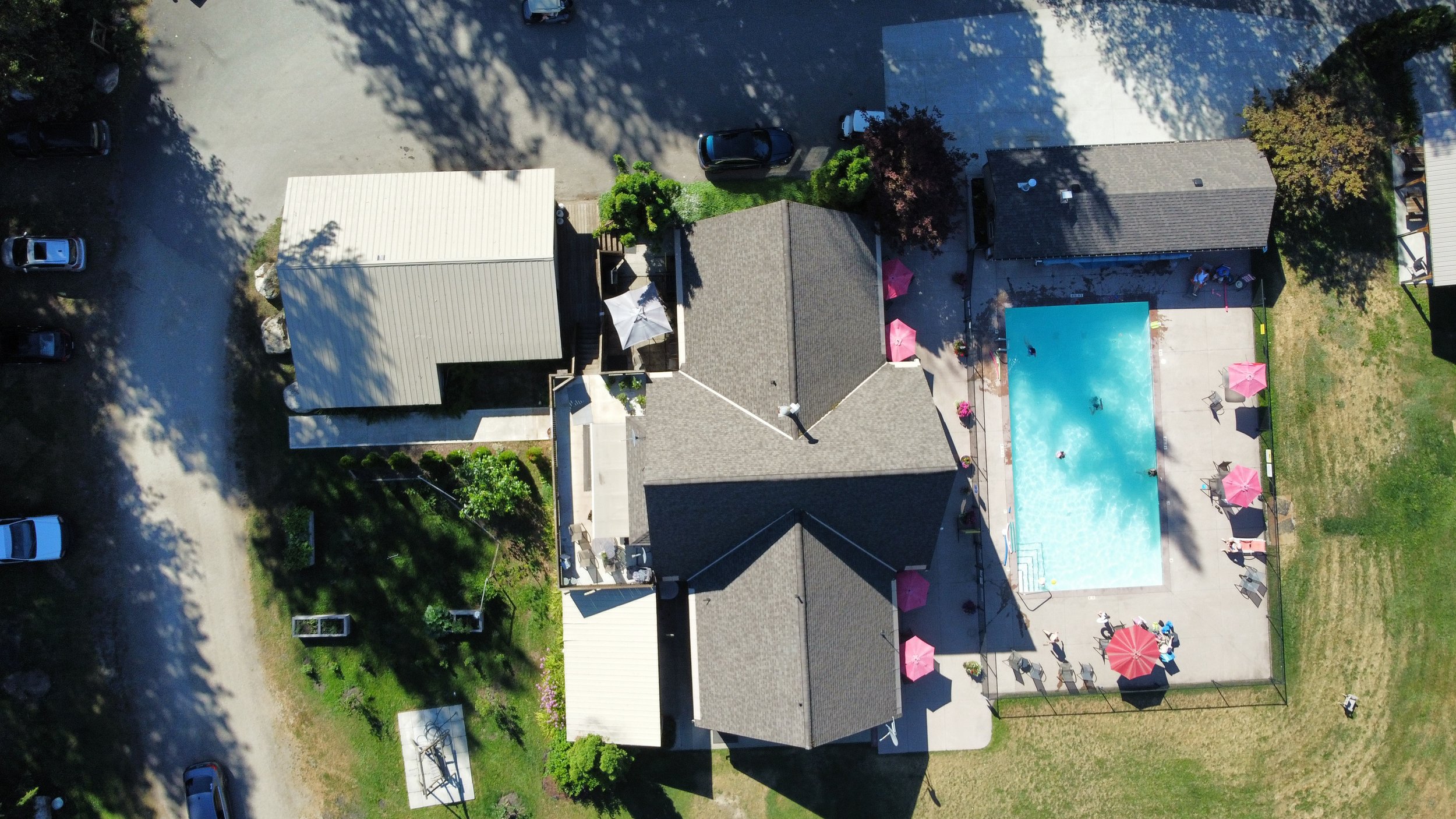 An aerial view of a residential backyard with a swimming pool, surrounded by pink umbrellas, chairs, and tables, with neighboring houses and a parking lot.