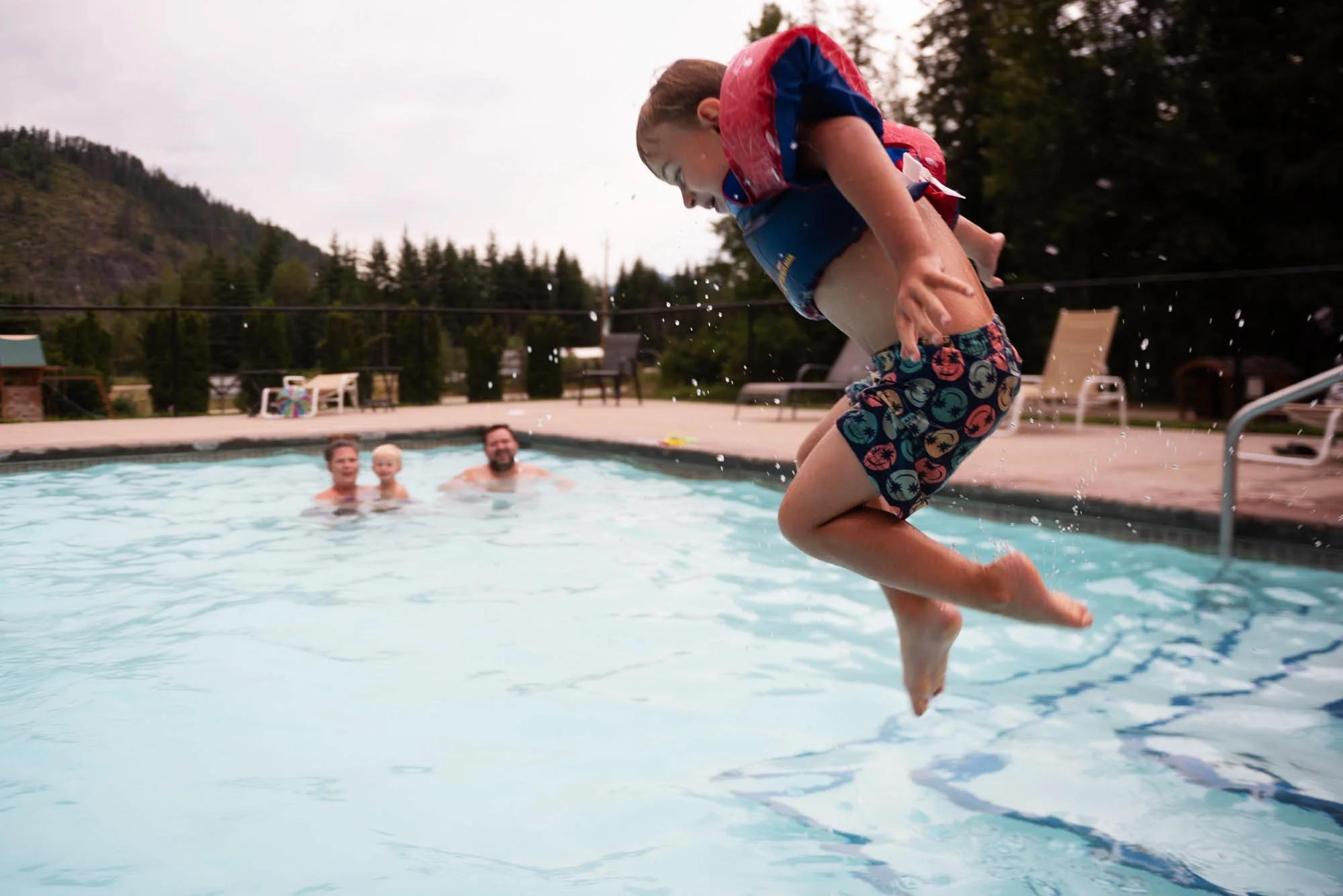A young boy jumping into a swimming pool with water splashing around, while adults watch in the background on a cloudy day.