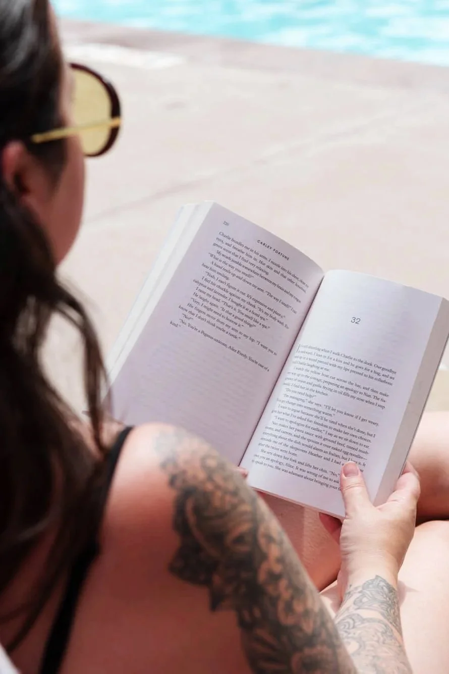 A woman with tattoos on her arm and wearing sunglasses reading a book while sitting beside a body of water at the beach.