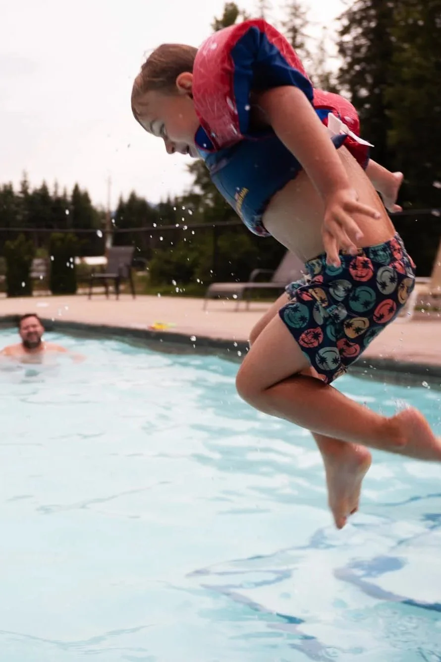 A young boy jumping into a swimming pool with water splashing around him, wearing a red life jacket and colorful swim trunks, while a man watches from the pool behind him.