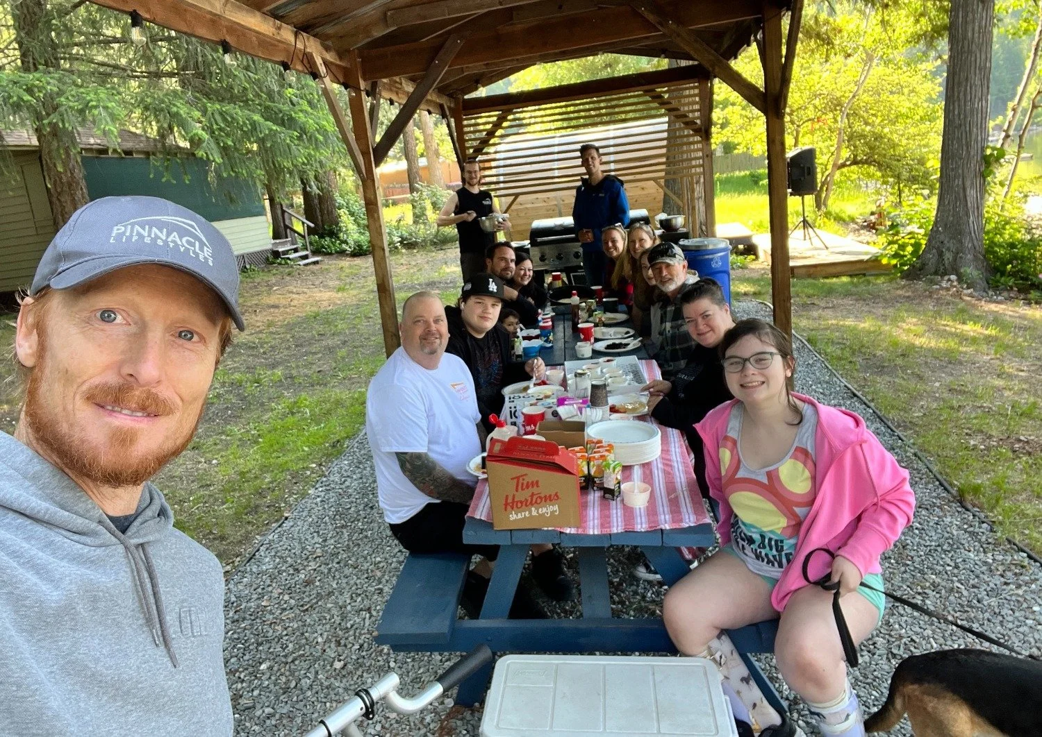 A group of people sitting at a long outdoor table enjoying a meal under a wooden shelter in a park or backyard. There is a man taking a selfie in the foreground, and others are smiling at the camera. The table has food and drinks, and some individuals are holding utensils or cups.