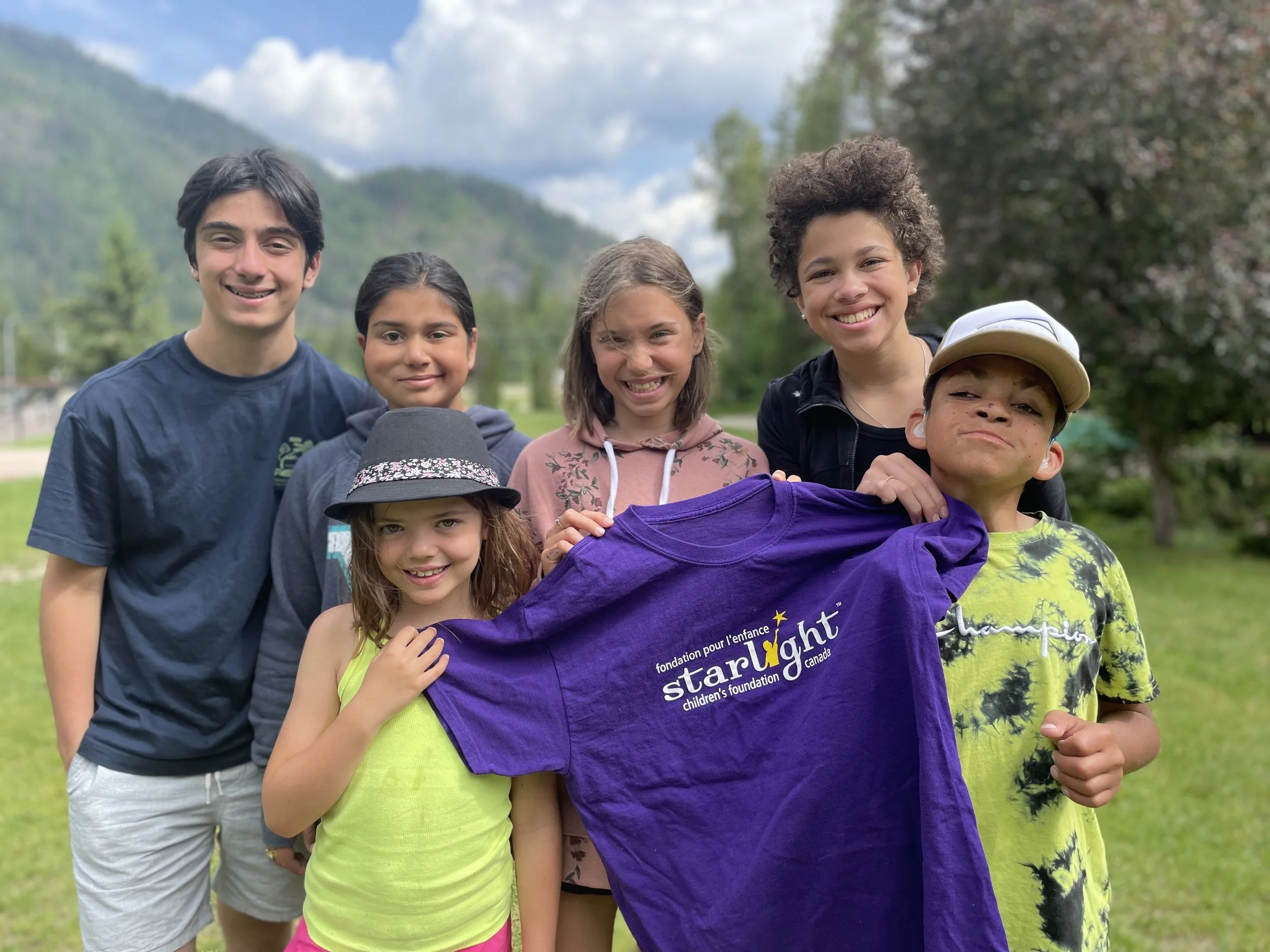A group of six children standing outdoors in a park with green grass and trees, smiling at the camera. One child is holding a purple T-shirt with the text 'starlight' and logos of a children's foundation, with mountains and cloudy sky in the background.