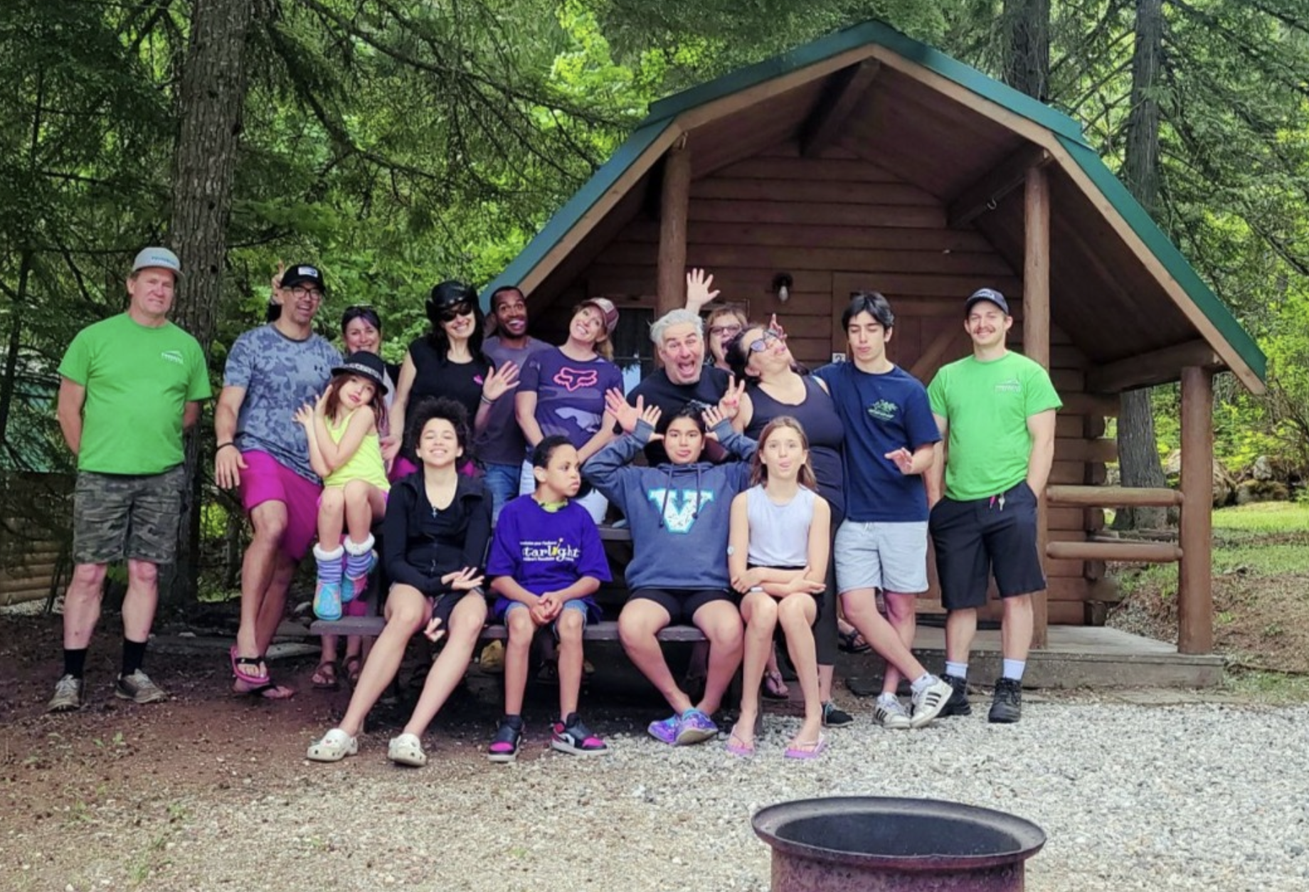 Group of children and adults smiling and posing in front of a wooden cabin in a forested area, some making playful gestures, and a fire pit visible in the foreground.