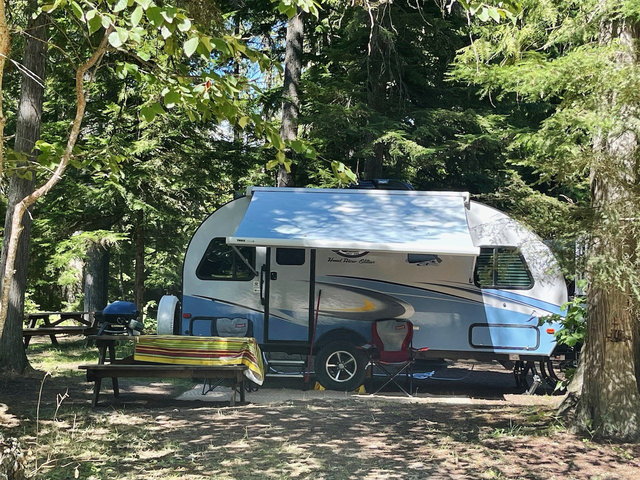 A travel trailer parked in a forest campsite with trees and shaded ground, accompanied by a picnic table with a striped tablecloth and camping chairs.