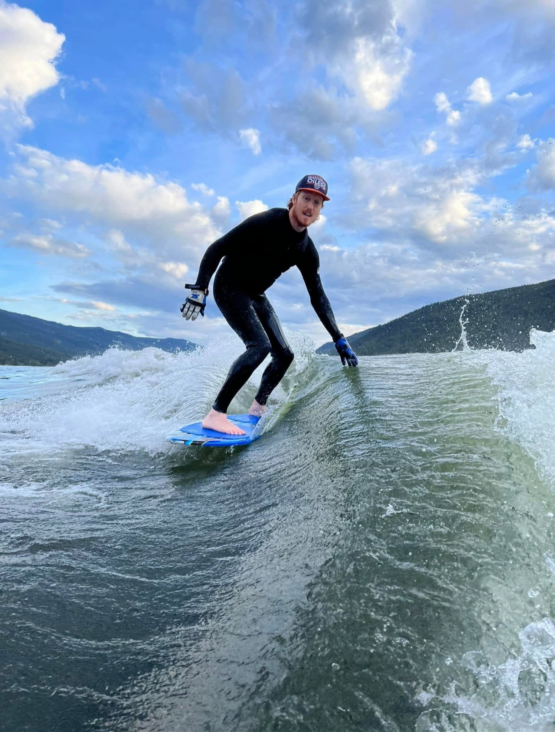 A man wearing a black wetsuit and a cap surfing on a small wave in a lake or ocean. The sky is partly cloudy with distant mountains in the background.