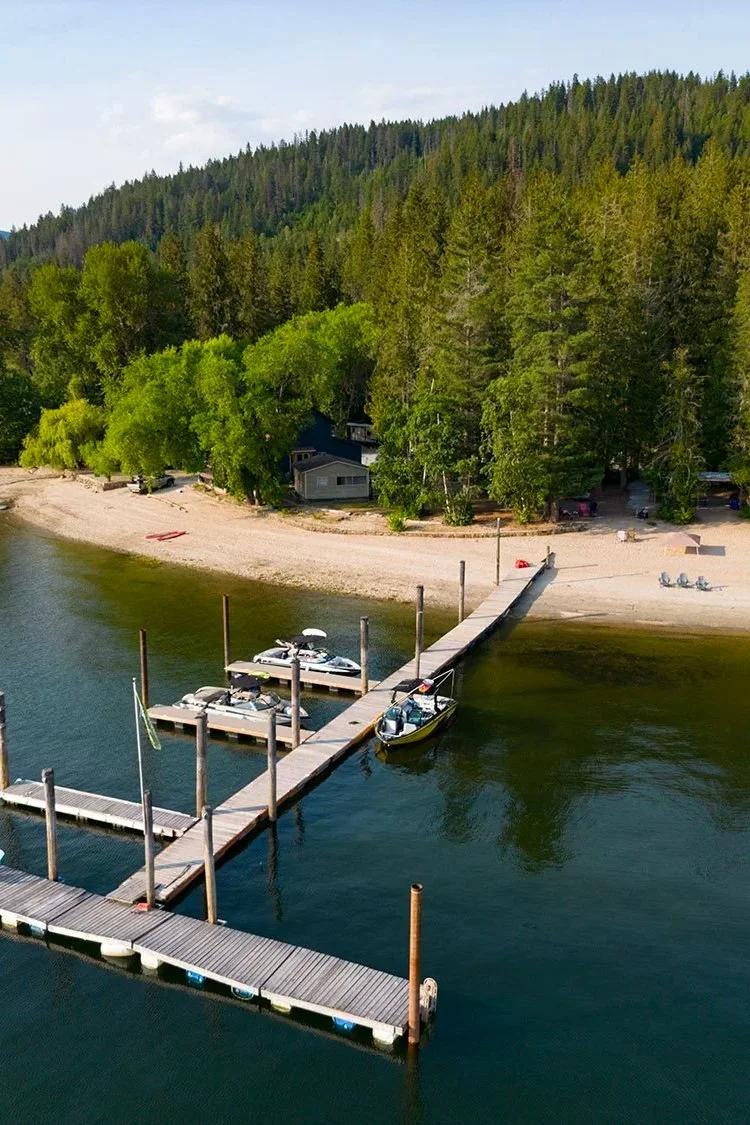 A boat dock extends into a body of water with several boats moored at the dock. Behind the dock, there is a sandy beach lined with green trees, and a wooded hillside in the background. Small structures and vehicles are visible among the trees.