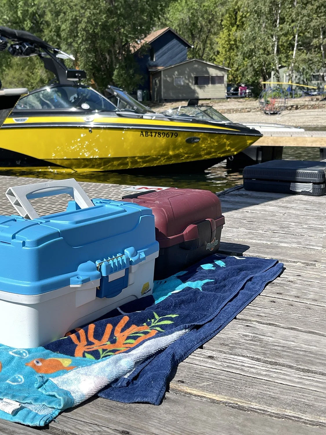 A yellow motorboat docked at a wooden pier on a lake, with various containers and a towel in the foreground, and houses and trees in the background.
