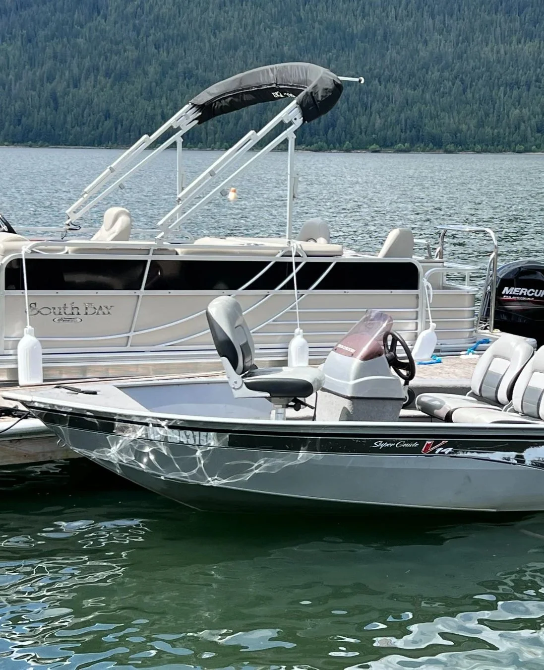 Two boats docked at a marina with water and trees in the background. One boat has seating and a dashboard, the other is a pontoon with a canopy.