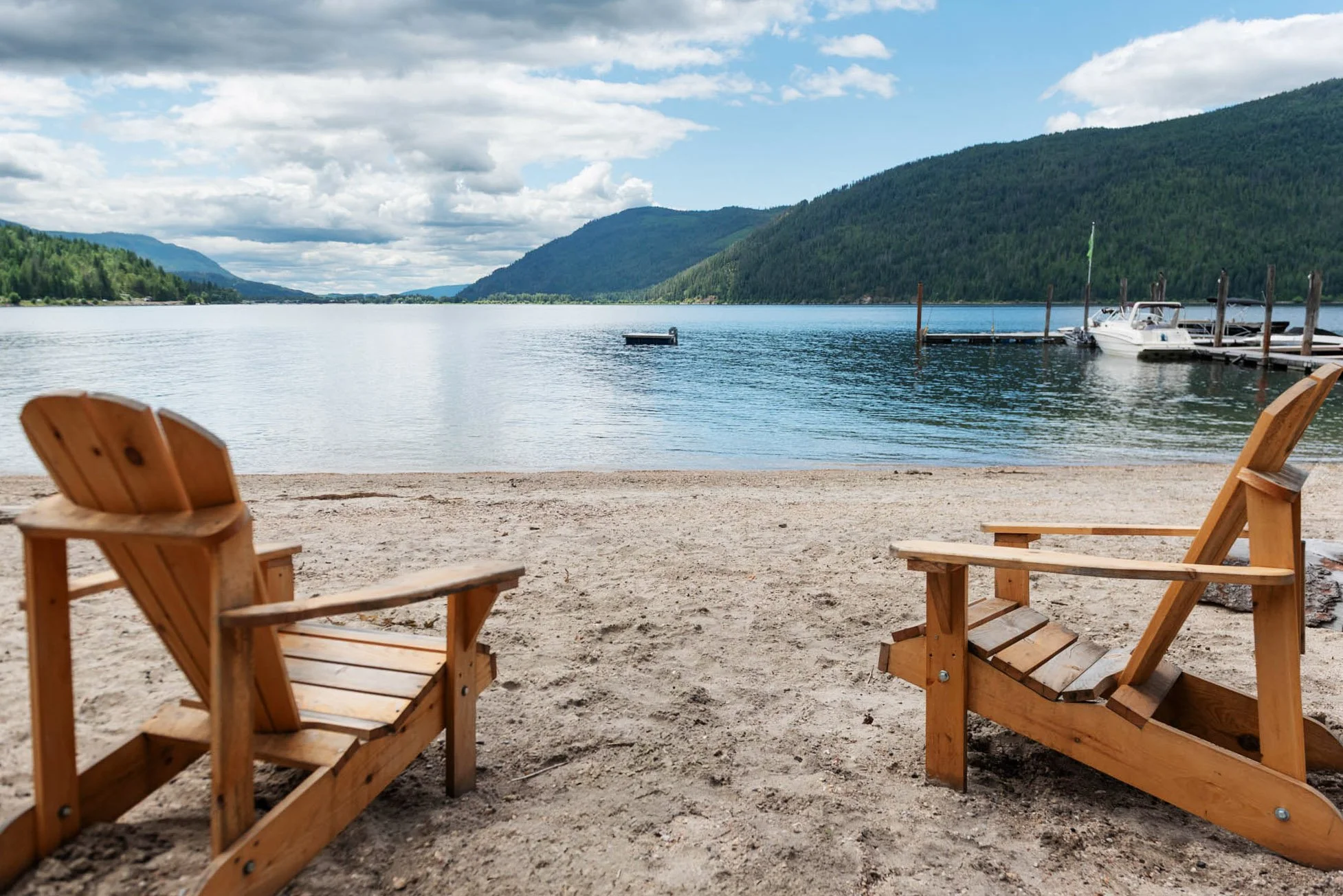 Beach scene with three wooden Adirondack chairs on sandy shore overlooking a lake with docks, boats, and green forested hills under a partly cloudy sky.