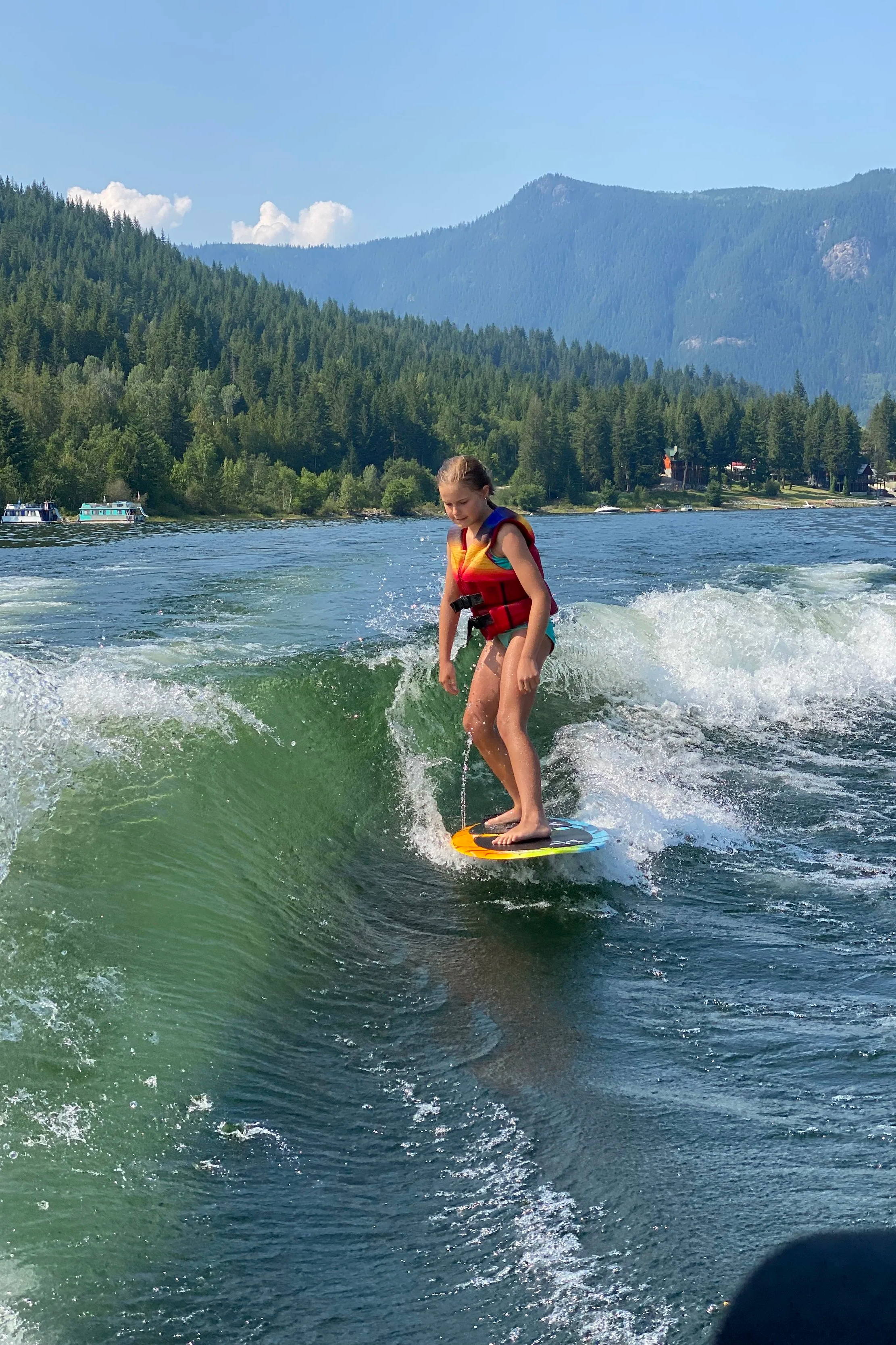 A young girl wearing a red life jacket surfing on a wave on a lake with a forested shoreline and mountains in the background.