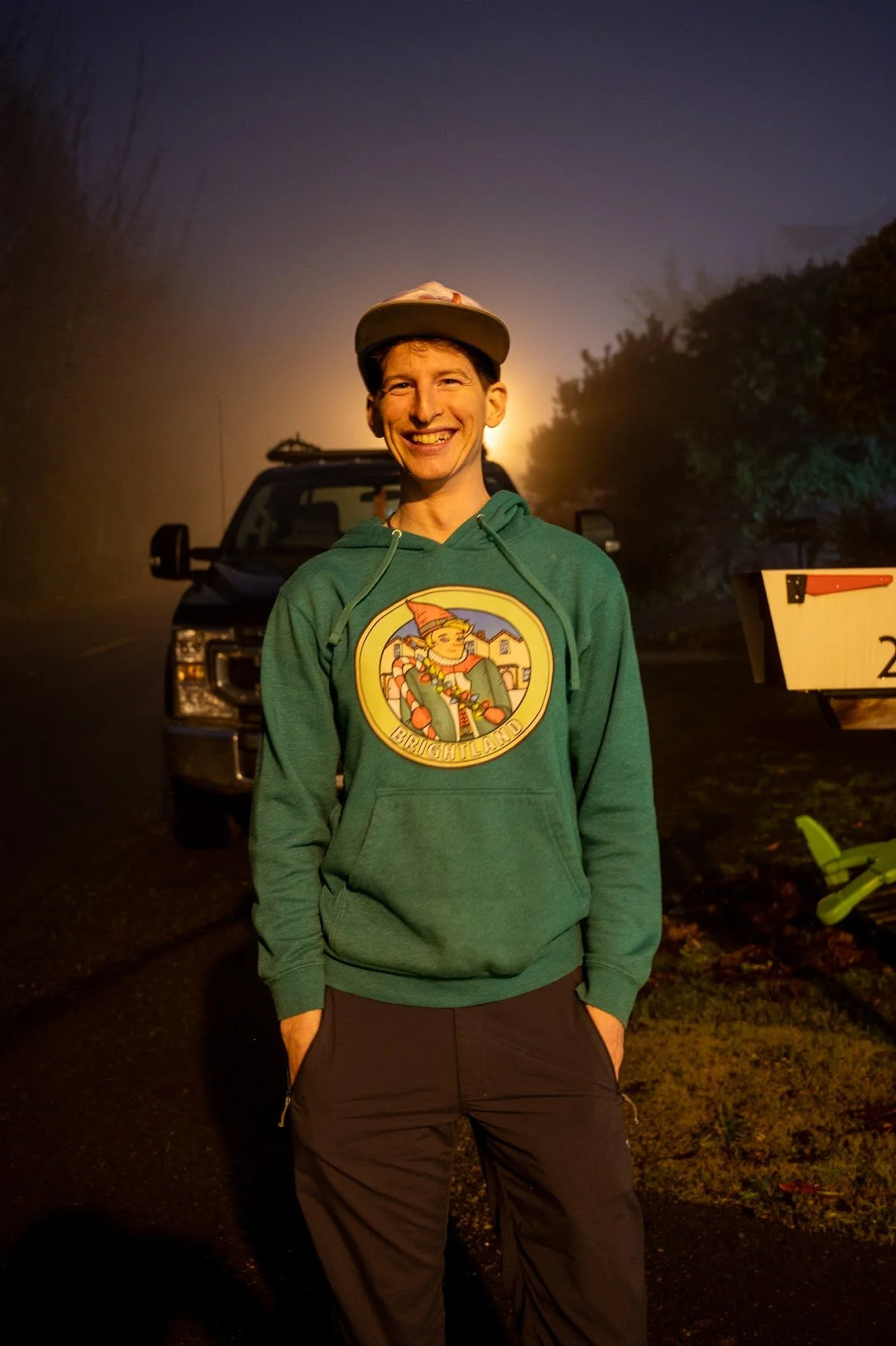 A smiling young man wearing a green hoodie with a colorful graphic and the text 'Brightland,' standing outdoors at night, with a vehicle and trees in the background.