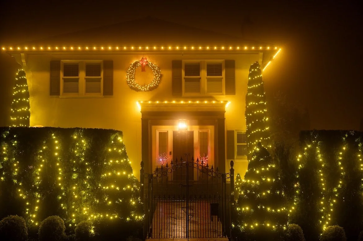 A two-story house decorated with Christmas lights, a wreath with a red bow on the front feature, and illuminated trees and bushes in the yard at night.