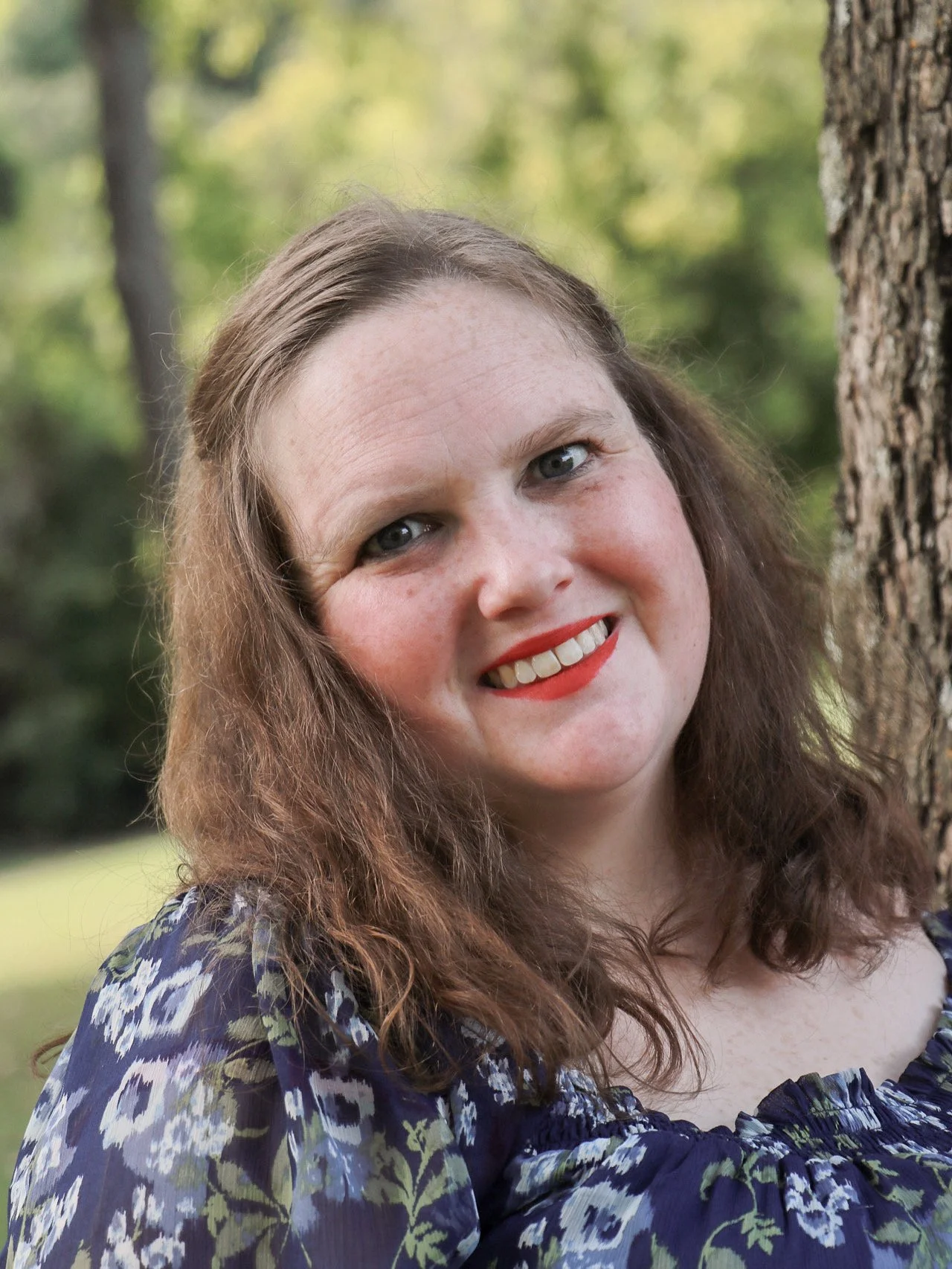 Smiling woman with long curly brown hair, wearing a blue floral dress, standing outdoors near a tree in a park or forest.