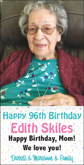 A smiling elderly woman with gray hair and glasses, wearing a turquoise top with white and purple floral patterns, a maroon cardigan, and a necklace, sitting in a chair.
