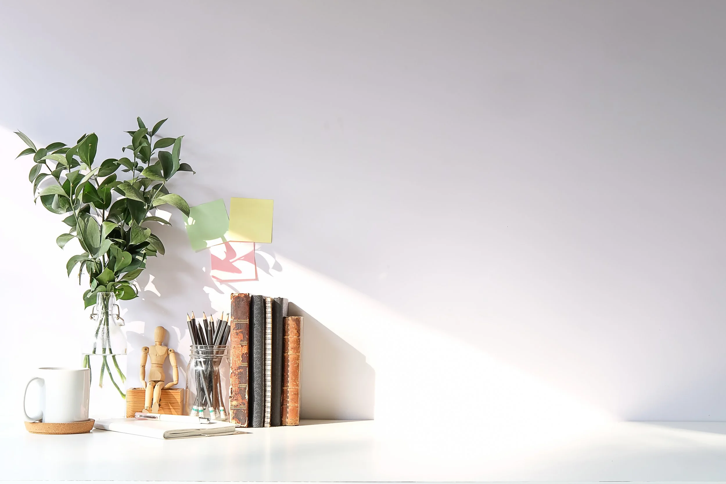 A minimalistic workspace scene with a white desk against a white wall. On the left, there is a glass vase with green leafy plant. To the right, there are three books with textured covers, a wooden mannequin, a transparent jar filled with pencils and pens, and a white mug on a tray. Sticky notes are attached to the wall behind the plant.