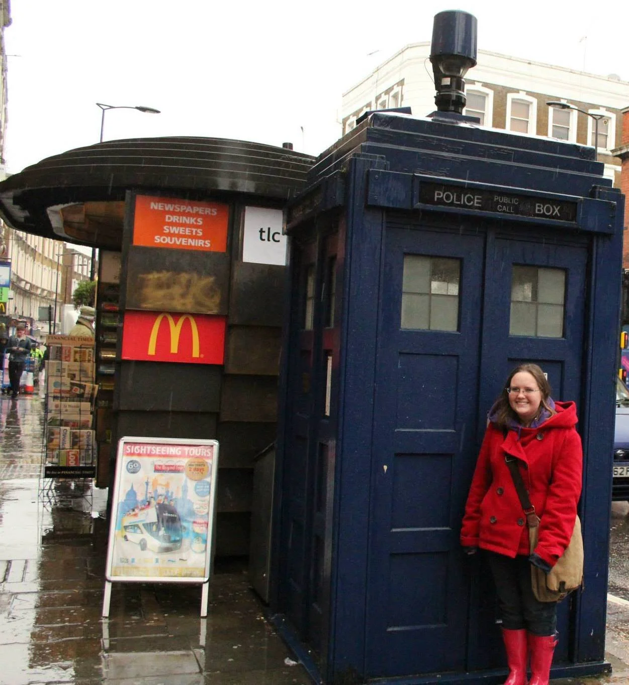 A woman in a red coat and rain boots standing next to a blue British police box on a rainy city street, with McDonald's signs and newspaper stand in the background.
