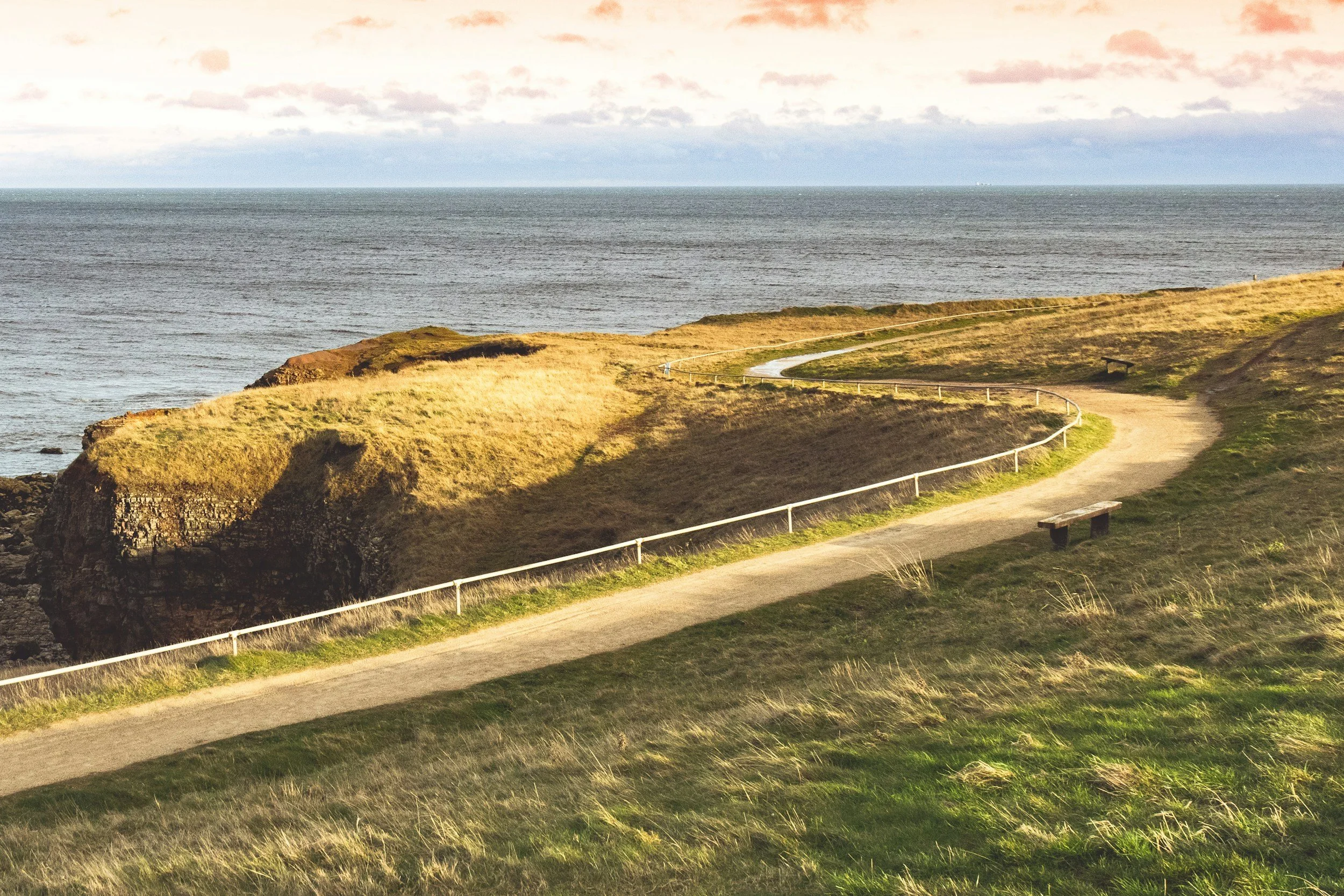 A coastal landscape showing a dirt path with a white railing, grassy hills, and the ocean in the background under a sky with clouds.
