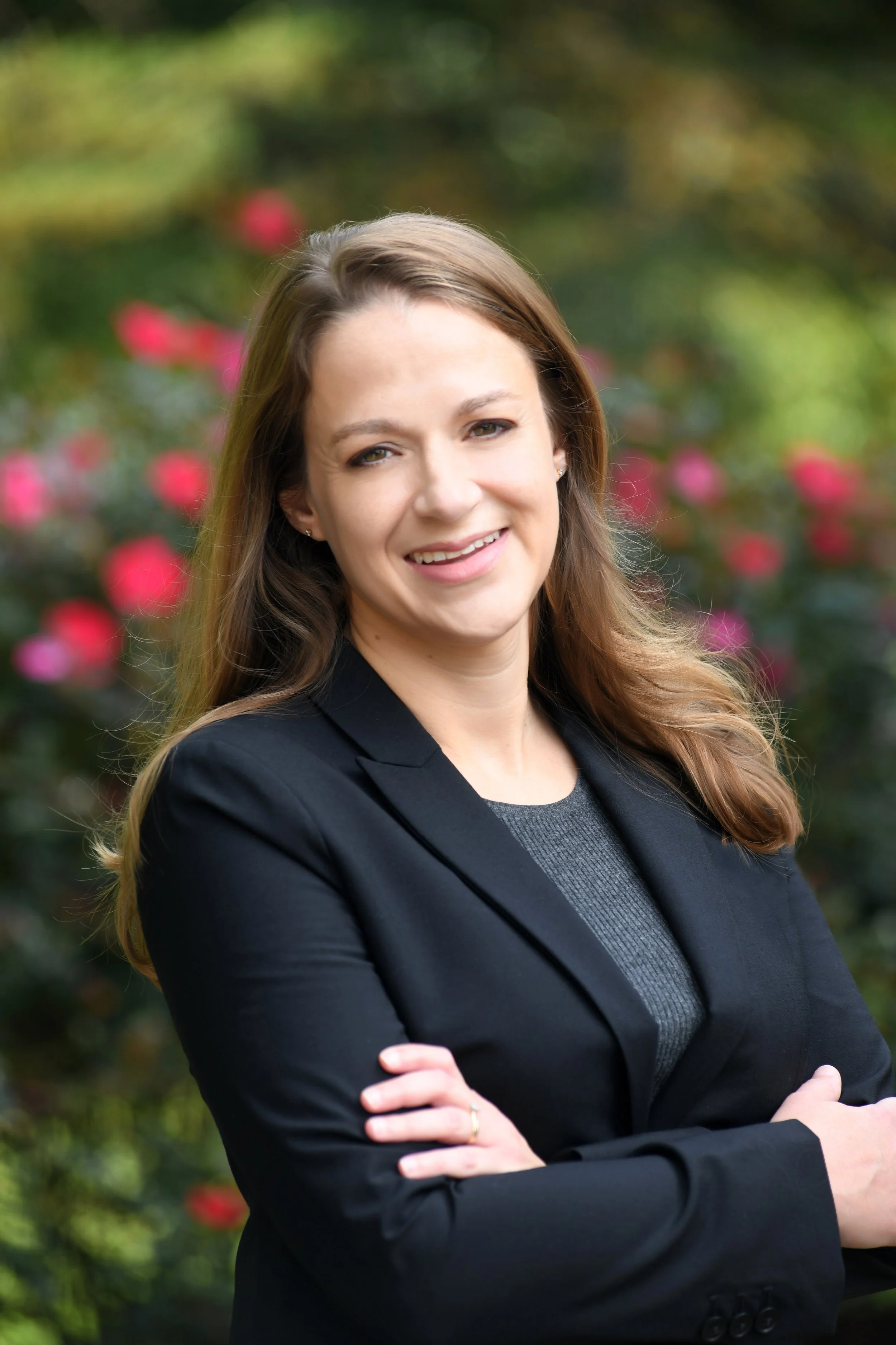 A woman with light brown hair smiling with cross arms, wearing a black blazer, outdoors with a blurred background of greenery and pink flowers.
