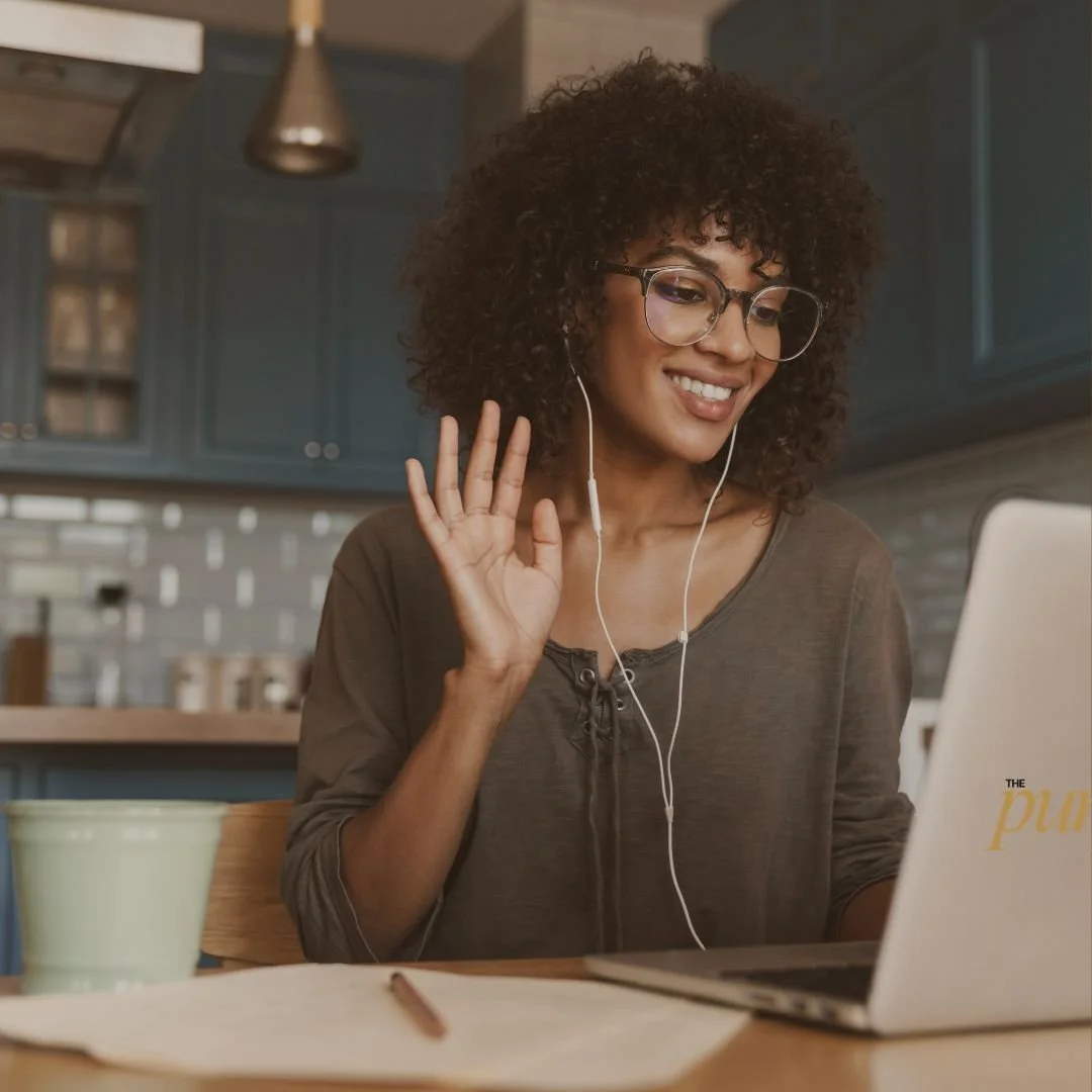 Woman with curly hair, glasses, and earphones smiling and waving at her laptop in a kitchen.