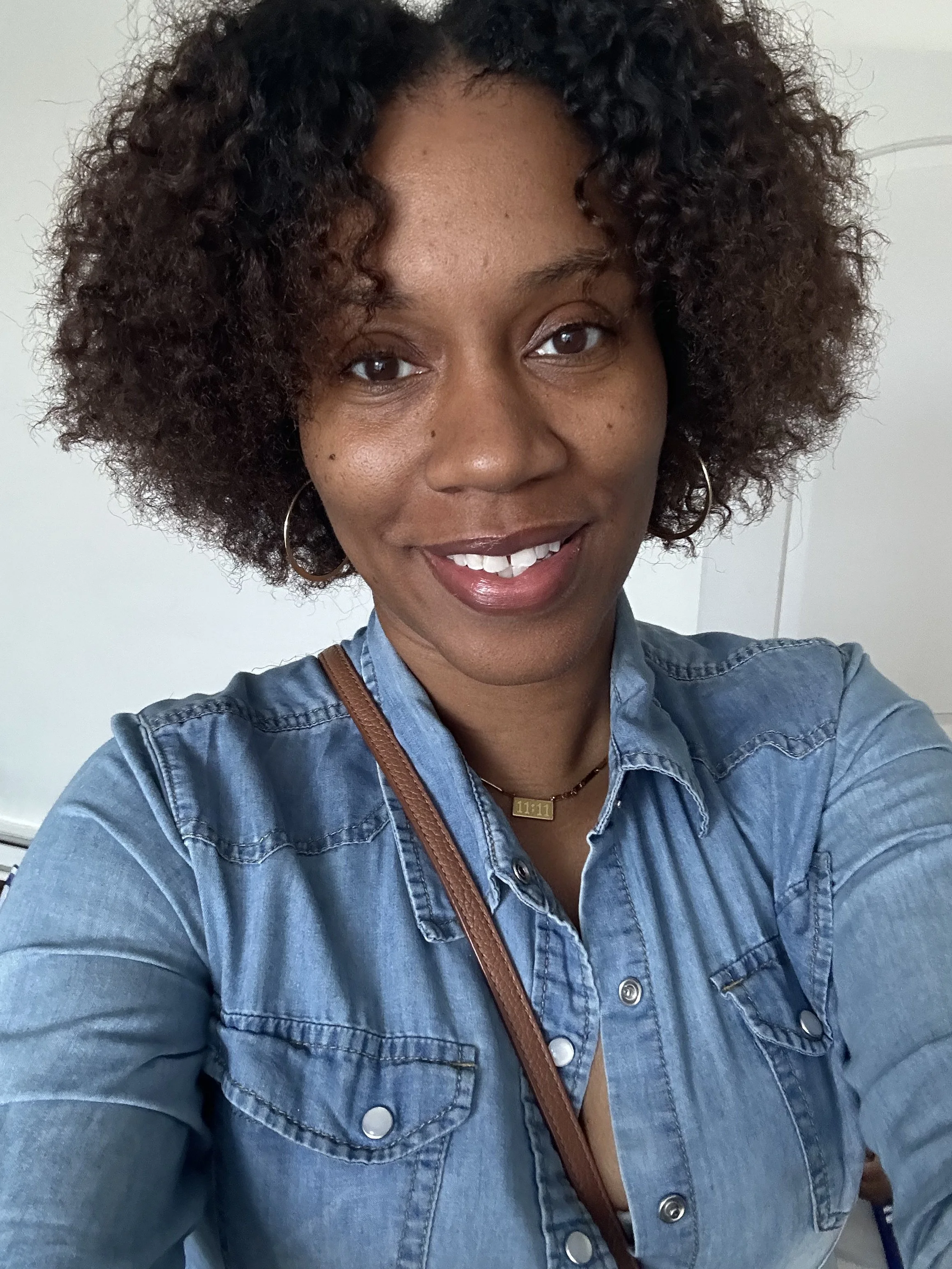 Close-up of a smiling woman with curly hair wearing a denim shirt, gold hoop earrings, a necklace with a rectangular pendant, and a brown strap bag.