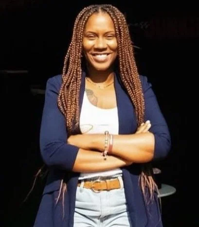 A smiling woman with braided hair, wearing a blue blazer over a white top and light jeans, standing with arms crossed against a dark background.
