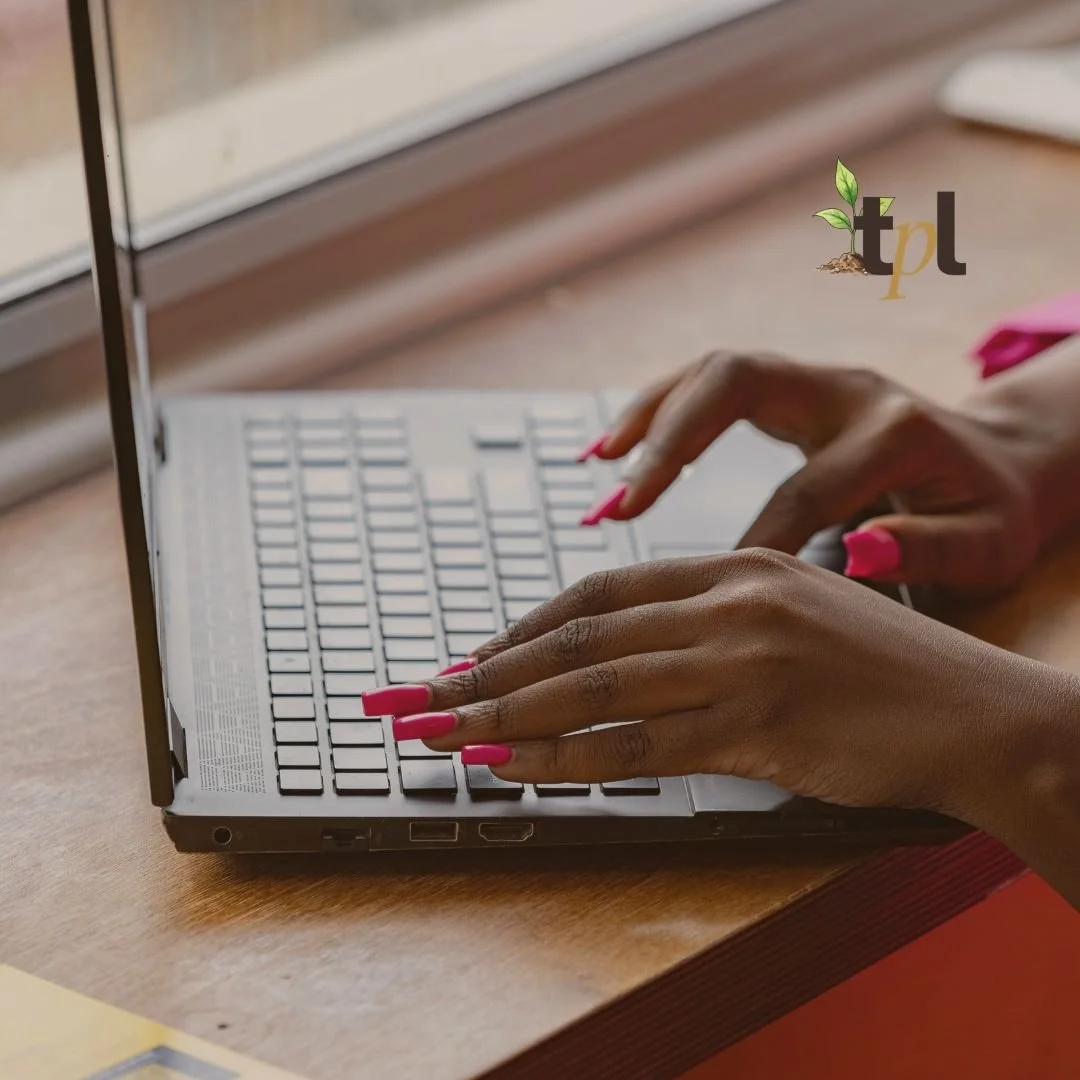 Close-up of hands with pink nails typing on a silver laptop keyboard on a wooden desk near a window.